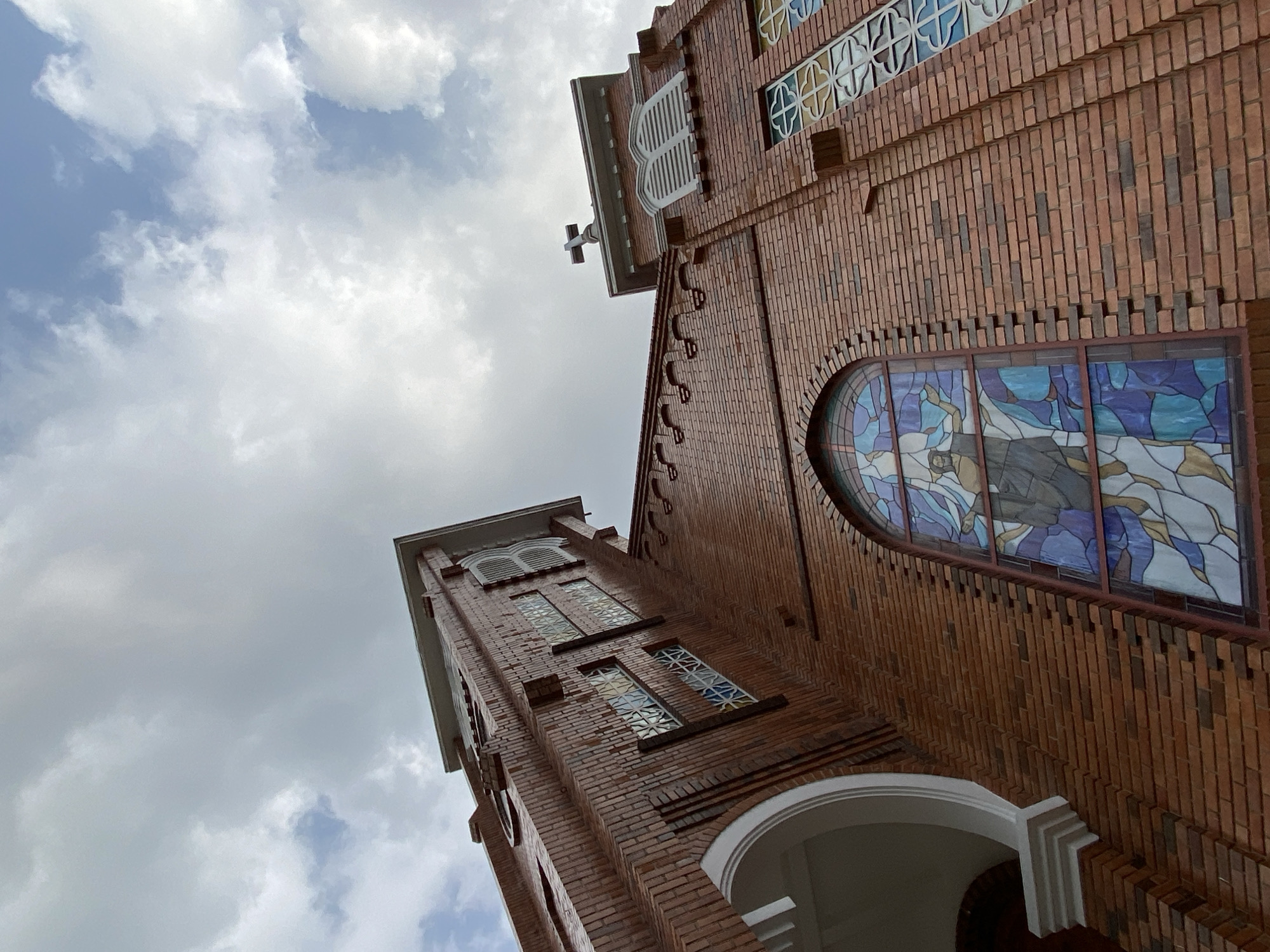 This low-angle shot captures the imposing facade of a red-brick building, likely a church, against a cloudy sky in Lâm Viên - Đà Lạt, Vietnam. The architecture features distinctive reddish-orange bricks laid in various patterns, creating a textured surface.
On the right, a prominent arched stained-glass window depicts figures in shades of blue, white, and brown, suggesting a religious scene. Further up, several smaller rectangular and square windows also feature intricate stained-glass patterns within white frames. To the left, a tall, tower-like section rises, topped by a white cross silhouetted against the sky. This tower also has arched window openings with white louvers and other patterned windows.
The sky is filled with large, soft white clouds, with occasional glimpses of pale blue sky, indicating a daytime setting. White arched architectural elements are visible at the base of the building, providing a contrasting detail against the brickwork. The perspective emphasizes the height and grandeur of the structure. No visible text is present in the image.