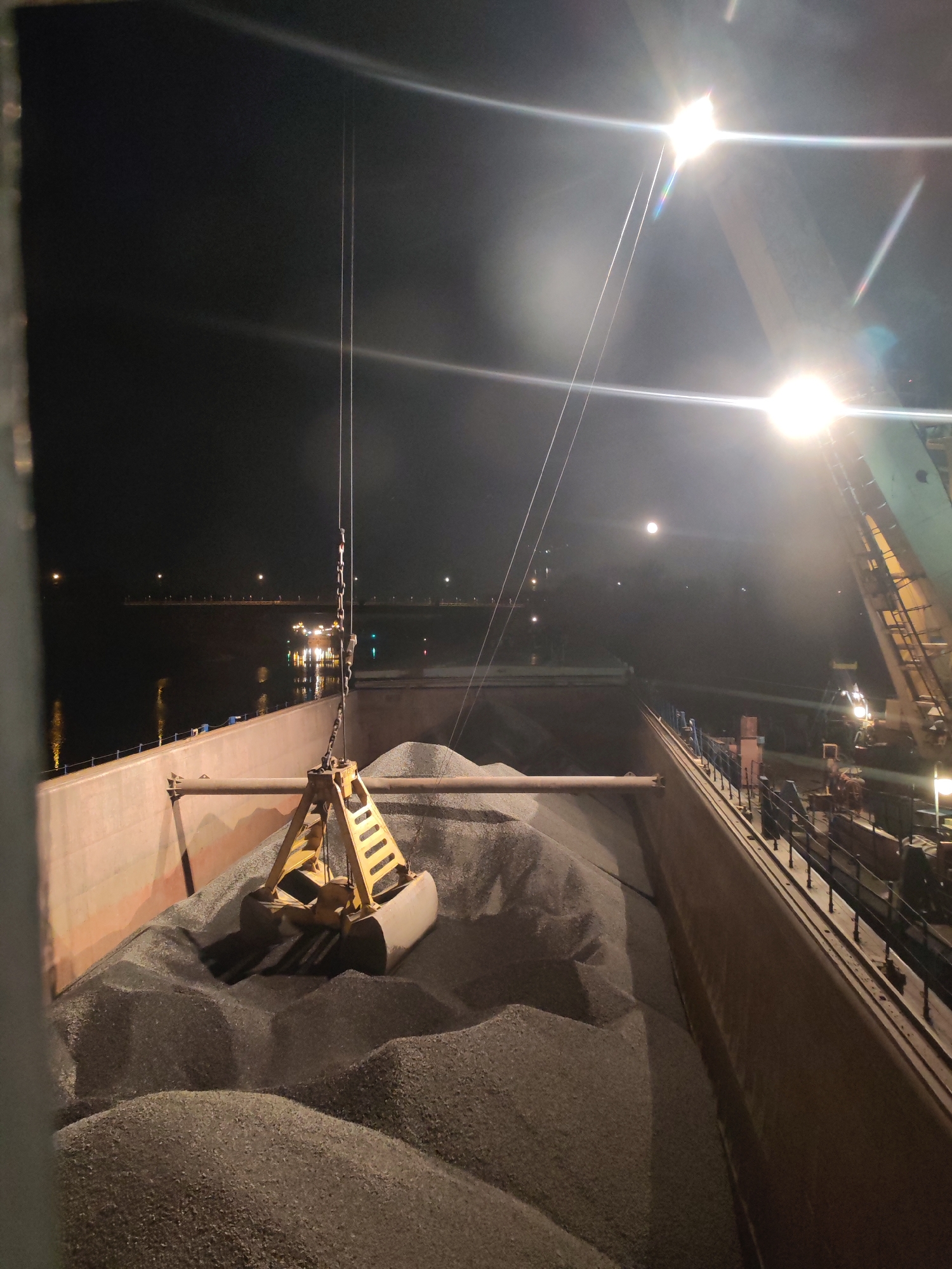 A nighttime shot shows a large, open-topped barge filled with gravel. A yellow industrial claw is lowered into the gravel. Bright lights illuminate the scene, creating glare and reflections. Beyond the barge is a body of water with lights reflecting on the surface. The scene is likely the Yakhroma river in Russia, where there is water flowing with gravel. There are no people visible in the image.