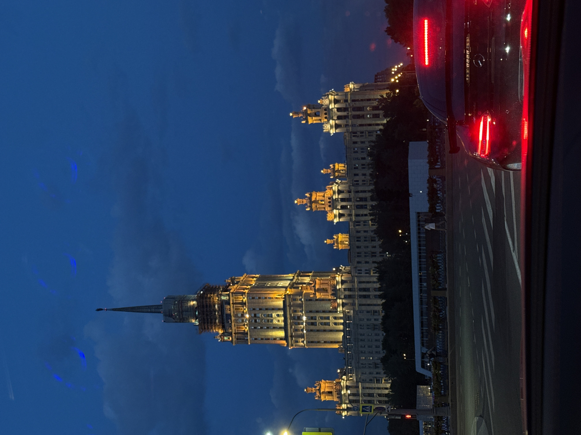 The image captures a night scene in Moscow, Russia, showcasing a magnificent illuminated building, likely one of the city's iconic Stalinist skyscrapers, against a dark blue, cloudy sky.
The prominent subject is a grand, multi-tiered building, possibly a Stalinist "Seven Sisters" skyscraper, standing tall on the left side of the frame. Its intricate architectural details, including cornices, decorative facades, and stacked sections, are highlighted by warm yellow artificial lighting. The building culminates in a distinctive, slender spire that pierces the dark sky. Small, subtle blue lights or reflections are visible in the clouds near the spire.
In the right foreground, the rear of a dark-colored vehicle is partially visible, with its bright red taillights and brake lights glowing intensely. This suggests the vehicle is either stopped or moving slowly on a street. A paved road with white lane markings is visible below the car, extending towards the bottom center of the image.
The sky is a deep, dark blue, indicating either late dusk or nighttime, with soft, visible clouds overhead. The artificial lighting on the building and the car's lights provide the primary illumination for the scene. Other indistinct buildings and trees are barely discernible in the background, further emphasizing the urban environment at night. No discernible text is visible in the image.