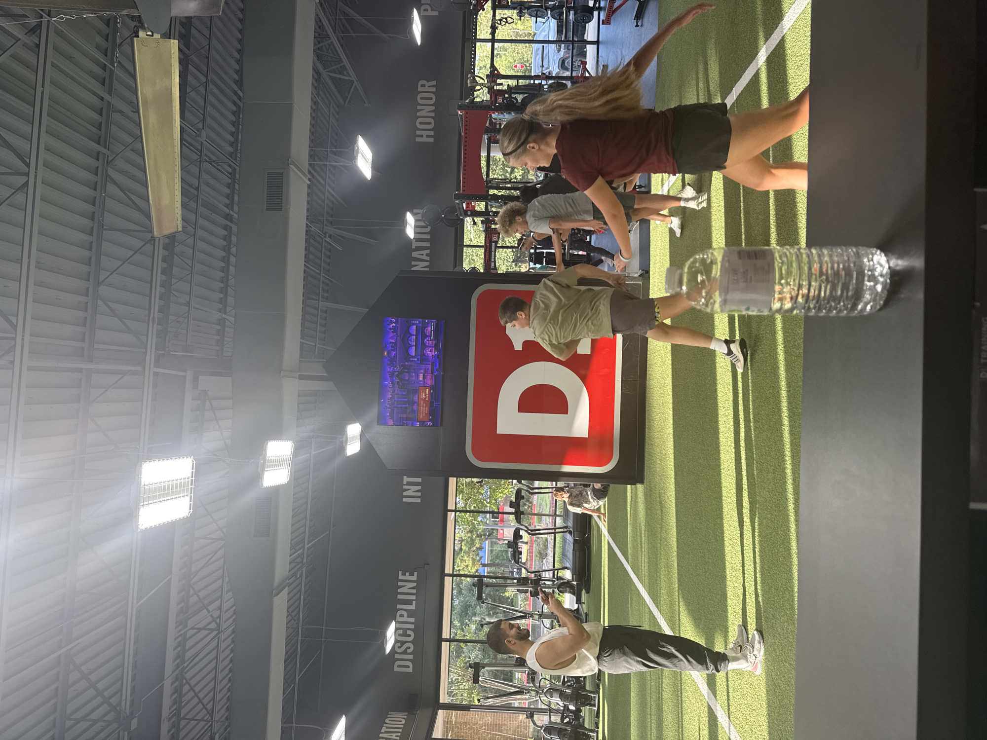 The image depicts the interior of a brightly lit gym, likely a D1 Training facility, given the prominent "D1" logo. The gym features a turf floor with white lane markings and various exercise equipment, including treadmills and weight racks, visible in the background. Natural light streams in from large windows, illuminating the space.
Several individuals are engaged in physical activity. In the foreground, a man wearing a white tank top and dark pants is standing and looking at his phone. Further back, a group of young people are participating in a workout. A blonde woman in a maroon shirt and black shorts is in mid-stride, her arms outstretched. To her left, a boy in a khaki shirt and grey shorts is also in motion. Another boy in a grey t-shirt and grey shorts is further back. A person in a patterned shirt is seated on the turf.
The gym's walls are adorned with motivational words like "DETERMINATION," "DISCIPLINE," "INTEGRITY," "NATION," and "HONOR." A large screen displays a colorful animated scene. A clear plastic water bottle sits on a dark surface in the foreground. The ceiling is high, with exposed metal beams and industrial lighting fixtures. The overall impression is a dynamic and active fitness environment.