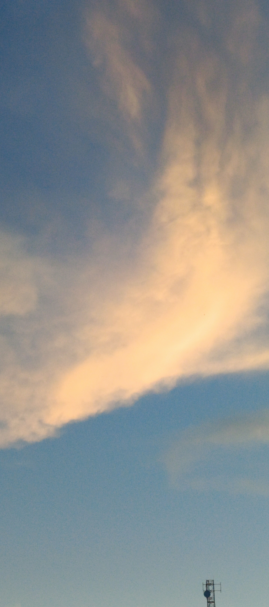 This image depicts a wide view of the sky with a prominent cloud formation illuminated by a warm, golden light, suggesting either sunrise or sunset. The sky transitions from a pale blue at the bottom to a deeper blue towards the top. The clouds are wispy and spread across the upper half of the frame, with the most intensely lit portion forming a curved band. In the lower right corner, a small portion of a telecommunications tower is visible against the clear sky. The scene is devoid of any human presence, and no specific urban landmarks of Sukamulia, Indonesia, are identifiable beyond the generic appearance of a telecommunications tower. The overall environment is that of an open sky, likely in a rural or suburban area where such towers are common.