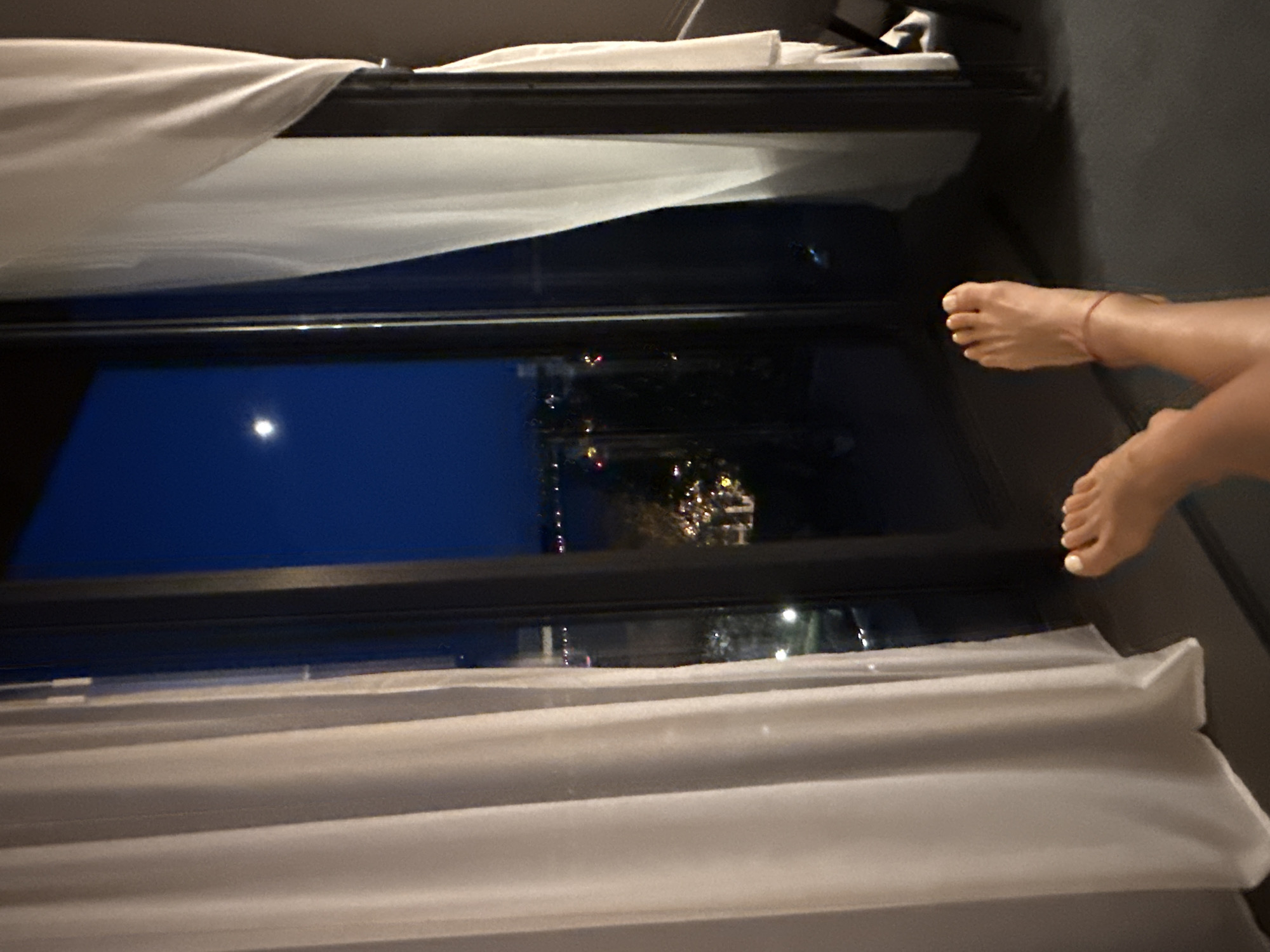 A person's bare feet are visible in the foreground, with a thin red string tied around one ankle. The feet are resting on a dark floor, and the legs extend upwards out of frame. Behind the feet is a large window that overlooks a city at night. The sky is a deep blue, with a bright moon visible. Below the sky, city lights twinkle amidst dark trees and buildings. White sheer curtains hang on either side of the window, partially obscuring the view. The scene appears to be from an apartment or hotel room in Kerch, Ukraine, offering a view of the cityscape at night.