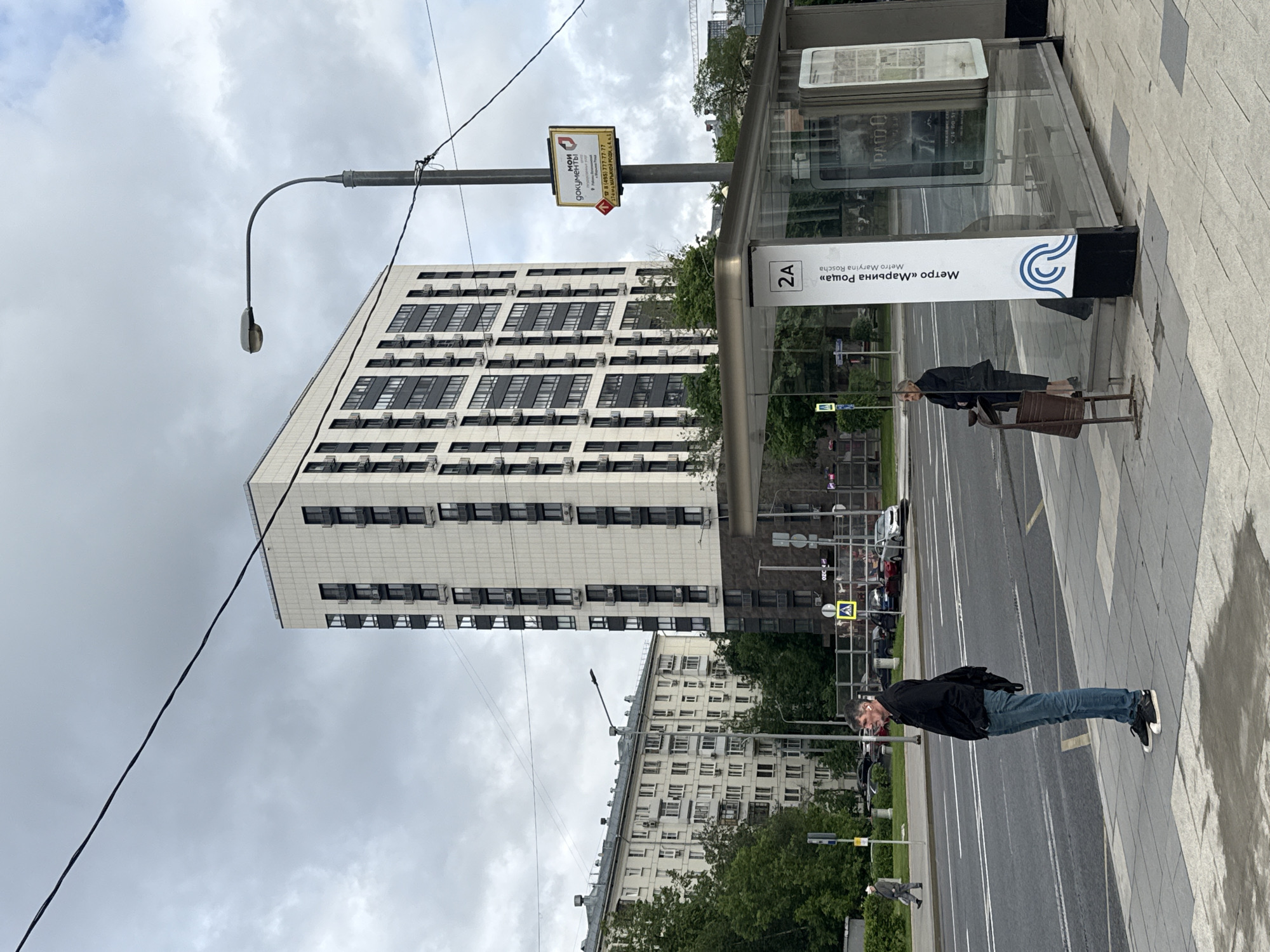 The image depicts a street scene in Moscow, Russia, featuring a modern building with numerous air conditioning units, a bus stop with signage for the "Maryina Roscha" metro station (Metro Maryina Roscha), and two individuals on the sidewalk. One person is standing near the bus stop, while the other is walking along the sidewalk. A sign advertising "Moi Dokumenty" (My Documents) is visible on a pole. The scene is set against a cloudy sky.