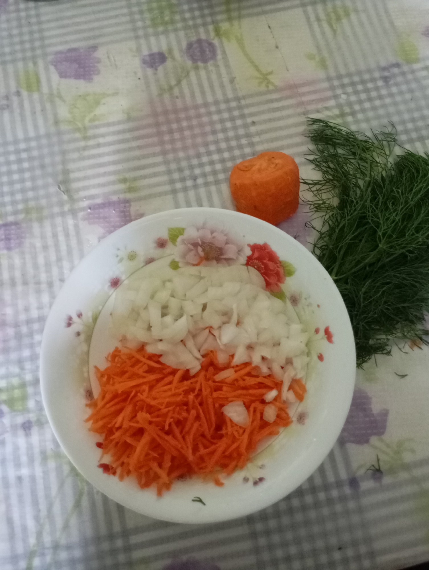 A white floral patterned plate holds chopped white onions and shredded carrots. A whole, peeled carrot cut into a round shape sits to the right of the plate, next to a bunch of fresh dill. The plate and vegetables are on a checkered tablecloth with purple floral accents. The scene is likely in a kitchen in Horishni Plavni, Ukraine, preparing for a meal.