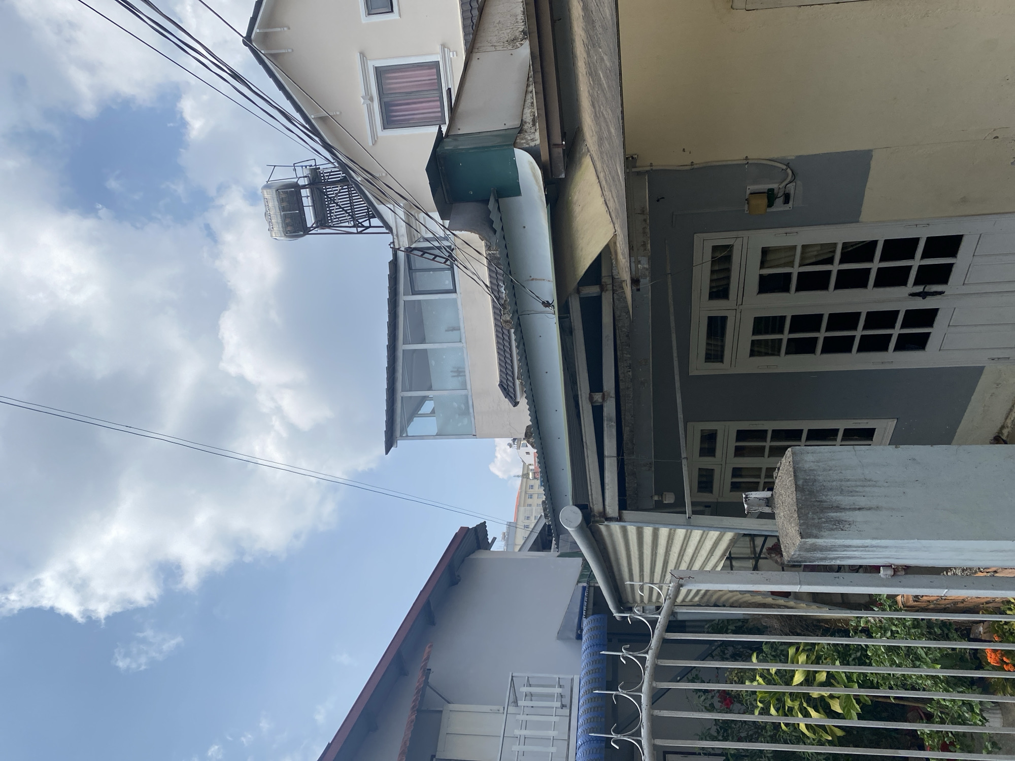 This image captures a densely built urban street scene in Lâm Viên - Đà Lạt, Vietnam, characterized by tightly packed, multi-story residential buildings viewed from a low angle, looking upwards towards a partially cloudy blue sky. The scene is illuminated by bright daylight, suggesting midday or early afternoon.
Several houses are visible, featuring various architectural details and colors including cream, yellow, grey, and white. Overhead, numerous black utility lines crisscross the sky, connecting different structures. A prominent stainless steel water tank, supported by a metal frame, is mounted on the rooftop of one of the buildings. Drainage pipes and gutters, some showing signs of wear and staining, run along the facades.
Architectural details include windows with multiple panes, some featuring decorative white frames. One window on an upper floor has a red curtain. On the right side of the image, a white, gridded door or large window is visible on a grey wall. In the foreground, a white metal fence or railing encloses an area where green foliage and some reddish-orange flowers are growing.
The overall impression is one of a lived-in, bustling urban environment with a focus on vertical structures and essential utilities, typical of a Vietnamese city. There is no discernible activity taking place, and no visible text is present in the frame.