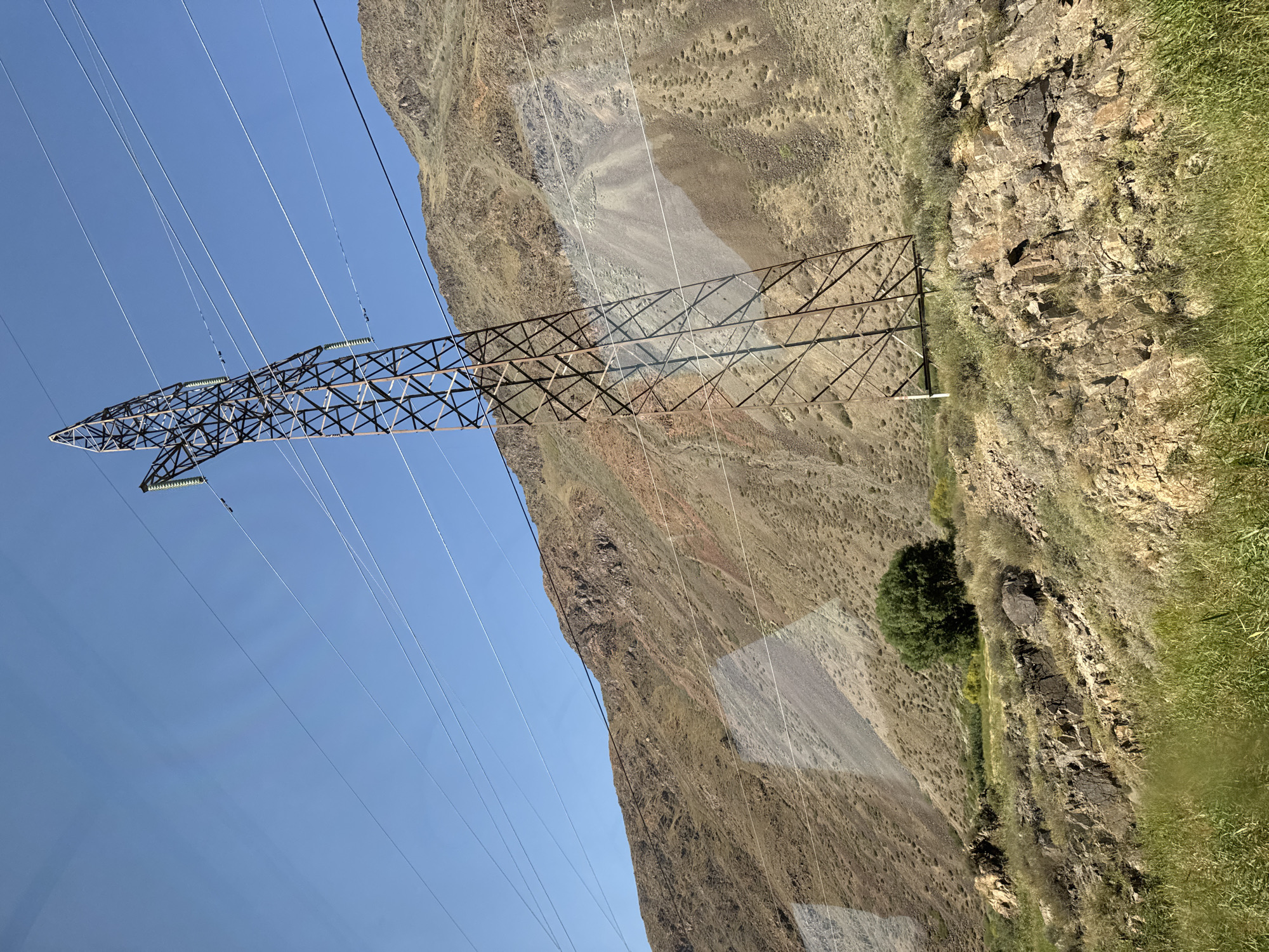 An outdoor medium shot captures a tall, metal electricity pylon dominating the foreground, leaning slightly to the left. The pylon is made of crisscrossed metal beams, weathered and rust-colored. It supports multiple power lines that stretch diagonally across the frame. In the background, a large, rugged mountain slopes upwards, its surface a mix of browns, grays, and reddish hues, indicating rocky and dry terrain with sparse vegetation. A single, bushy green tree stands on the lower slope of the mountain, to the left of the pylon's base. The sky above is a clear, bright blue. The scene appears to be in a natural, mountainous landscape, possibly near a rural or remote area, and is described as being in the city of Unknown, Kyrgyzstan.