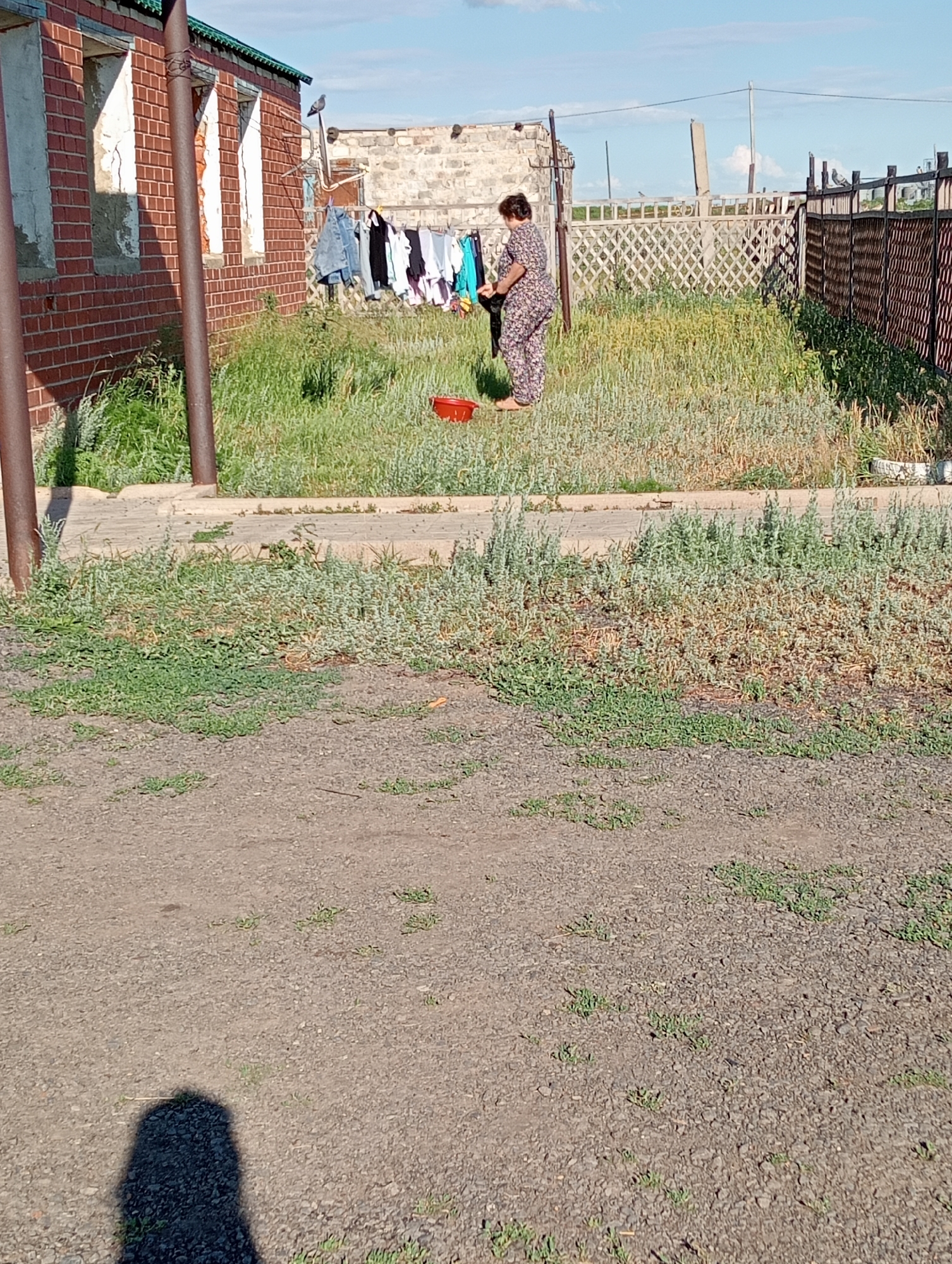 A woman in a patterned dress is hanging laundry on a clothesline in a rural setting. She is standing in front of a brick building with unfinished window openings, and the clothesline is strung between poles. Various items of clothing, including jeans, shirts, and what appear to be towels, are drying on the line. A red bucket sits on the ground near the clothesline. The scene is outdoors, with abundant green grass and some dry, wispy vegetation. A decorative lattice fence and a darker wooden fence are visible in the background. The sky is blue with some scattered clouds, suggesting a pleasant day. The sun appears to be casting shadows, indicating it is likely daytime. A shadow of a person is visible in the foreground. The overall impression is one of a humble, everyday activity taking place in a simple, rural environment. There is no visible text in the image.