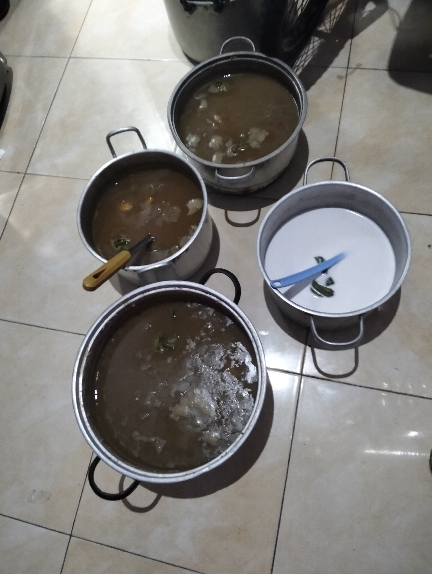 Four cooking pots are placed on a tiled floor. The pots are made of metal and have handles. One pot contains a white liquid with green leaves. Another pot contains a brownish liquid with chunks of food and a wooden utensil. A third pot also contains a brownish liquid with chunks of food and green leaves. The fourth pot is partially visible and also appears to contain a brownish liquid with food. The floor is tiled with light-colored square tiles. In the background, a portion of a black appliance with a metal rim is visible, possibly a stove or another cooking device. The lighting in the scene is dim, with shadows cast by the pots and the unseen objects in the background. This scene appears to be a domestic kitchen setting, likely in a residential area within the City of Medan, Indonesia.