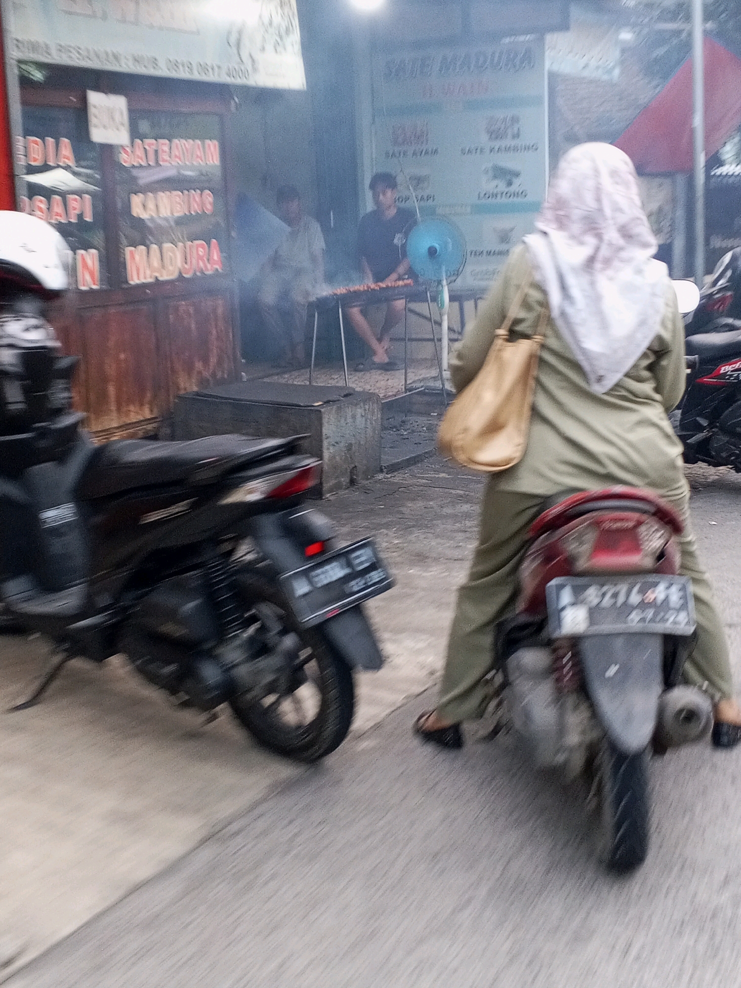 The image captures a street scene in Ciruas, Indonesia, likely at a food stall specializing in satay. In the foreground, a dark-colored motorcycle is parked, its rear partially visible. To the right, another motorcycle is also parked, with a person wearing a green hijab and green clothing seated on it, holding a tan shoulder bag. This person is facing away from the camera.
The background reveals a food stall with several signs. One prominent sign reads "SATE MADURA" and lists "SATE AYAM," "SATE KAMBING," and "LONTONG." Another sign, partially obscured by smoke, displays "SATE AYAM MADURA." Several people are visible near the stall, including one man tending to skewers over a grill, and another man sitting down. A blue fan is also present. The environment appears to be an outdoor setting, with some smoke emanating from the grill, suggesting cooking activities. The ground is paved with asphalt. The overall impression is one of a bustling street food vendor operation.