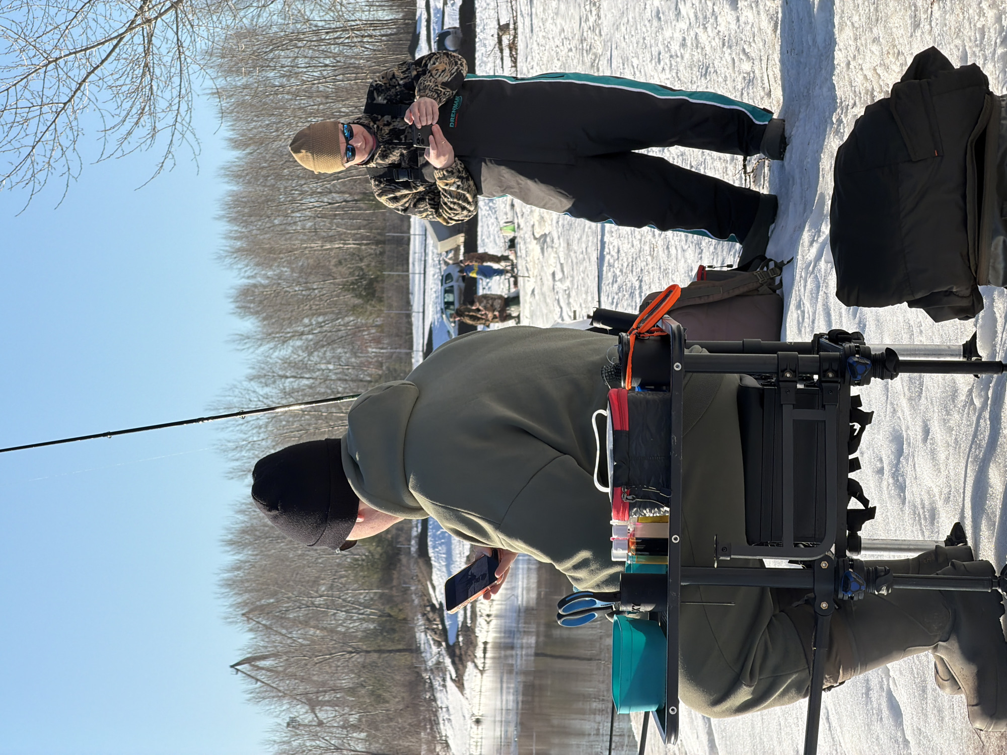 The image shows two individuals on a snowy terrain in what appears to be a rural setting. One person, wearing a khaki beanie, sunglasses, a camouflage jacket, and dark overalls with a turquoise stripe down the side, is standing and making a hand gesture towards the camera. A second individual, wearing a dark beanie and an olive green jacket, is seated on a black fishing seat and looking at a cell phone. A fishing rod is visible above the seated person. The scene contains fishing equipment attached to the seat. Snow covers the ground, and trees line the background under a clear blue sky. The presence of fishing equipment and a river visible in the middle of the image indicates a waterside location, but there are no specific indicators to place the image in Chaikovsky, Russia. There are other individuals in the background.