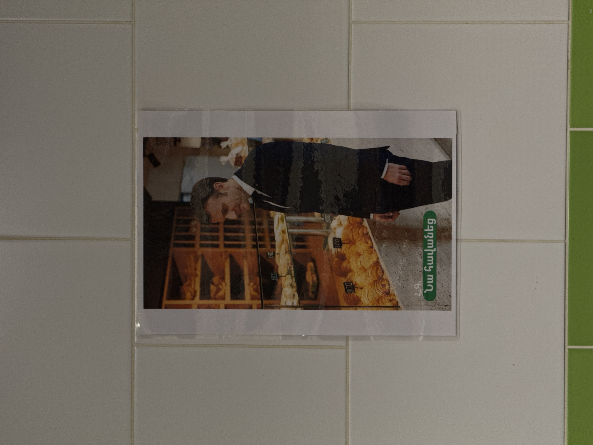 The image shows a poster affixed to a tiled wall. The poster appears to be an advertisement in Armenian, displaying a person in a suit and a bakery display case with priced goods. It is implied to be located in Yerevan, Armenia.