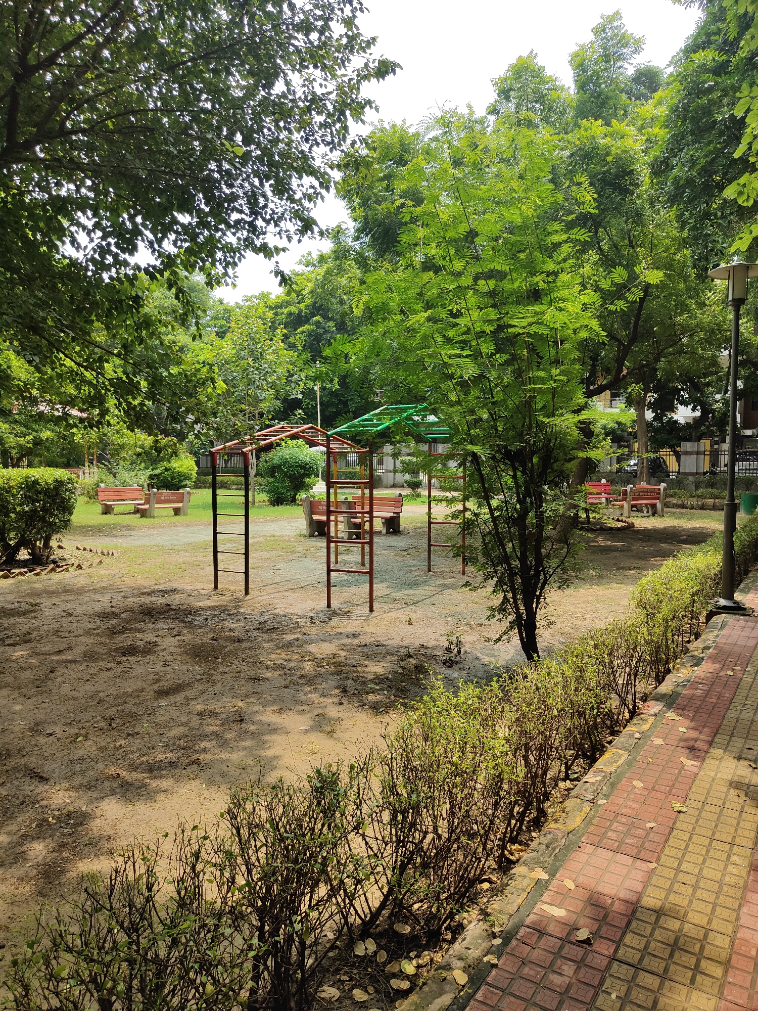 The image depicts a public park in Gurgaon, India, likely a residential area given the presence of buildings in the background. The park features a children's play structure with red and green canopies, red benches with concrete bases, and a sandy ground area. A paved walkway with red and yellow tiles runs along the right side of the image, bordered by manicured green bushes. Lush green trees and foliage are abundant throughout the scene, suggesting a well-maintained outdoor space. The lighting indicates it is daytime, with shadows cast by the trees on the ground, suggesting dappled sunlight. No people are visible in the frame.
