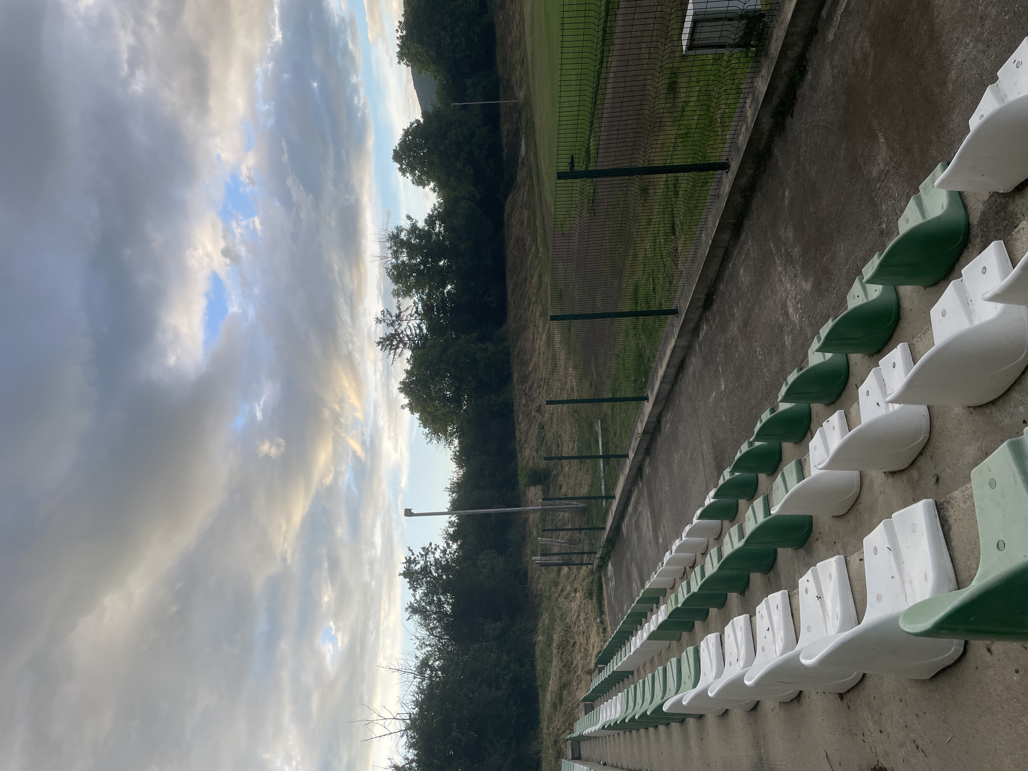 This image depicts an empty stadium or sports ground in Ihtiman, Bulgaria, captured during what appears to be late afternoon or early evening. The scene is dominated by rows of unoccupied spectator seats, alternating in white and green, running diagonally across the lower right of the frame on a concrete stand. The concrete surface shows signs of wear.
A dark green mesh fence separates the seating area from the field. Beyond the fence, there's a grassy expanse, which transitions from what looks like a well-maintained green playing surface closer to the fence, to more overgrown and dry grass further away. A dense line of dark green trees forms a natural backdrop behind the field, with one tall, slender pole visible among them. Faint outlines of hills or mountains can be seen on the horizon above the treeline.
The sky occupies the upper portion of the image, presenting a mix of weather conditions. Large, grey clouds are prominent, but there are also patches of clear blue sky peeking through. Some clouds are illuminated with a soft, warm, yellowish light, suggesting the low angle of the sun, indicative of either sunrise or sunset. No people or animals are visible, contributing to a sense of quietude. There is no visible text in the image. The overall impression is one of stillness, perhaps between sporting events or awaiting an upcoming one.