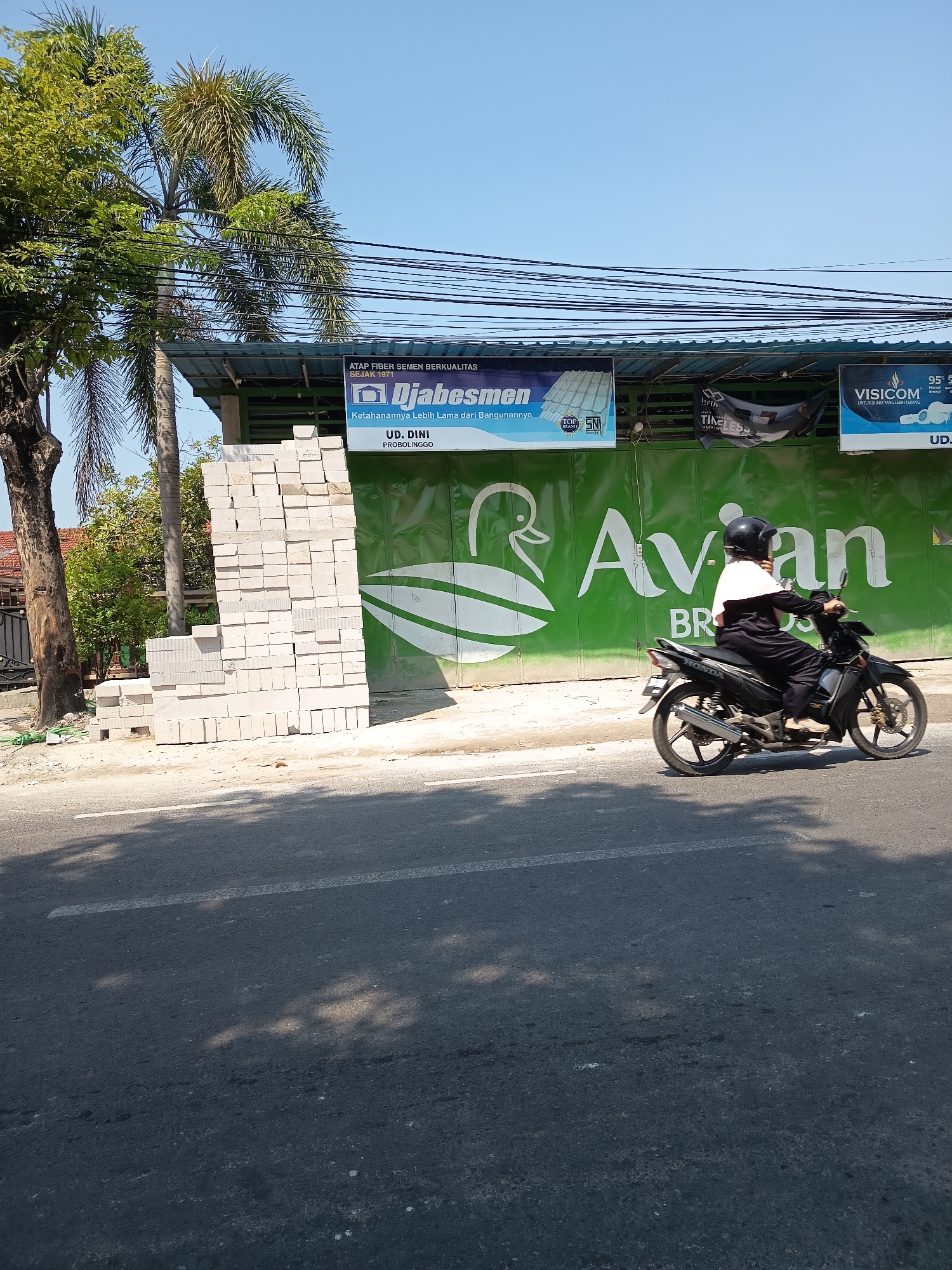 A motorcycle with two people, a driver and a passenger, both wearing helmets, is riding on a paved road in Kandangjati Kulon, Indonesia. The motorcycle is black and silver. The driver is wearing a black outfit and a white cape over their shoulders. The passenger is also wearing black. They are on the right side of the road, moving away from the viewer. Behind them, a large green building with a white logo of a bird and the text "Avian BR" is visible. To the left of the building, there is a stack of white bricks. A tall palm tree with green fronds is to the far left of the image. Power lines are strung across the top of the scene, against a clear blue sky. The road has a white line marking. Shadows of trees are cast on the road.