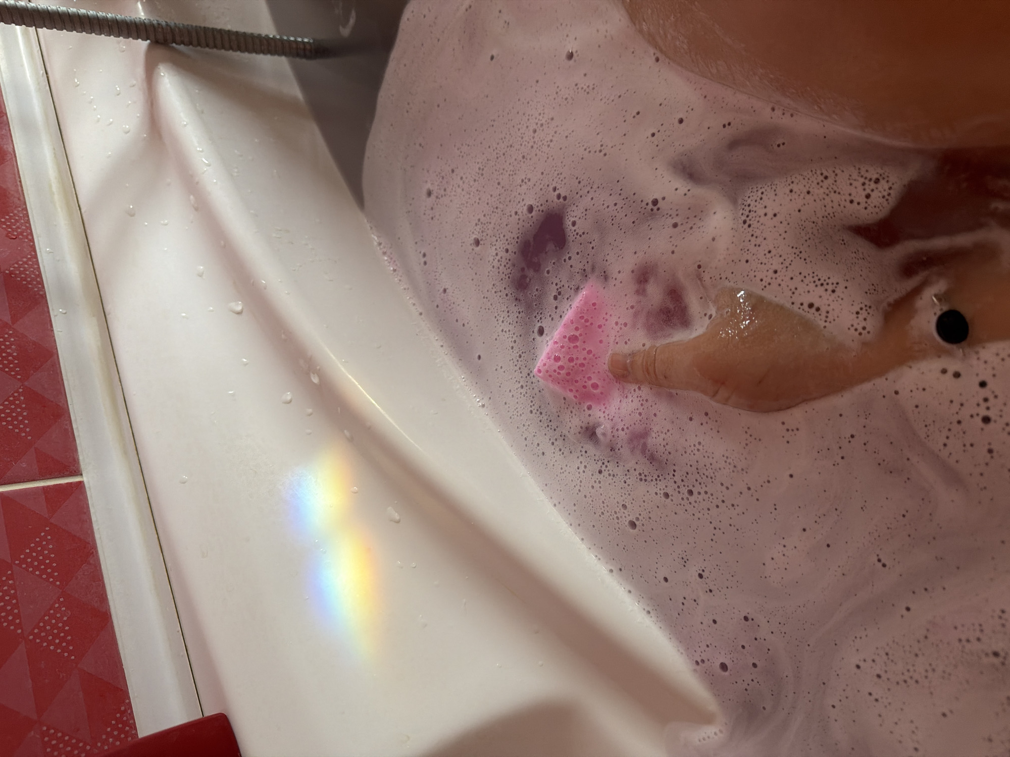 The image depicts a person in a bathtub filled with pink bubble bath. A pink sponge is being held by a hand within the bubbly water. The side of the white tub is visible, reflecting a rainbow of light. The bathtub appears to be in a bathroom, with red and white patterned tiles visible on the edge. The location is Большая Шильна, Russia.