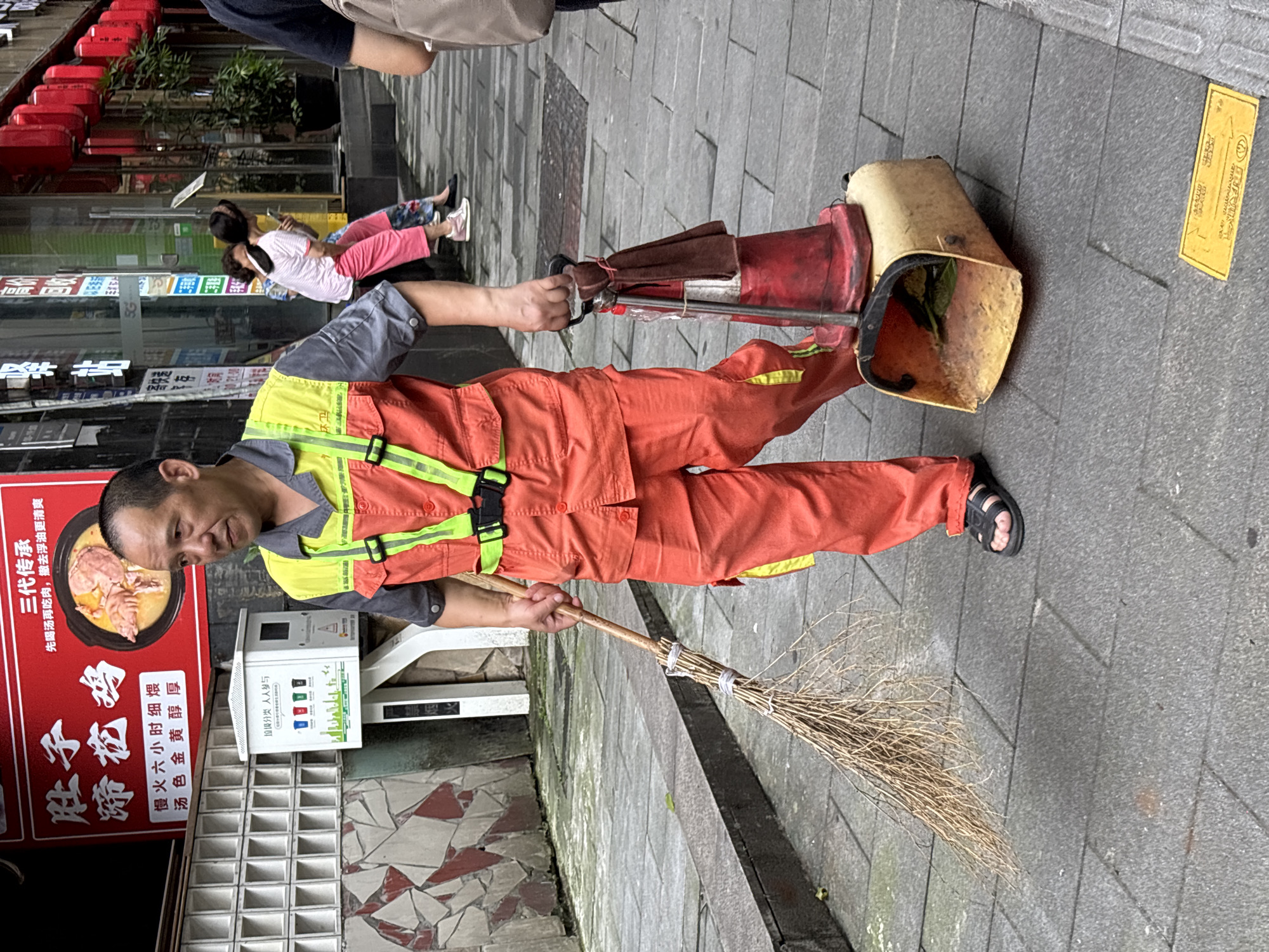 A middle-aged East Asian man, appearing to be a street sweeper, stands on a gray tiled sidewalk in Yuzhong District, China. He wears an orange uniform with a fluorescent yellow vest and reflective straps. He is holding a traditional broom in his left hand and a trash receptacle with a red cone attached on its handle in his right hand. The trash receptacle contains some green leaves and debris. In the background, there is a red sign with Chinese characters and an image of food, a white recycling bin, and two women walking away. The environment appears to be an urban street with buildings and hanging red lanterns visible.