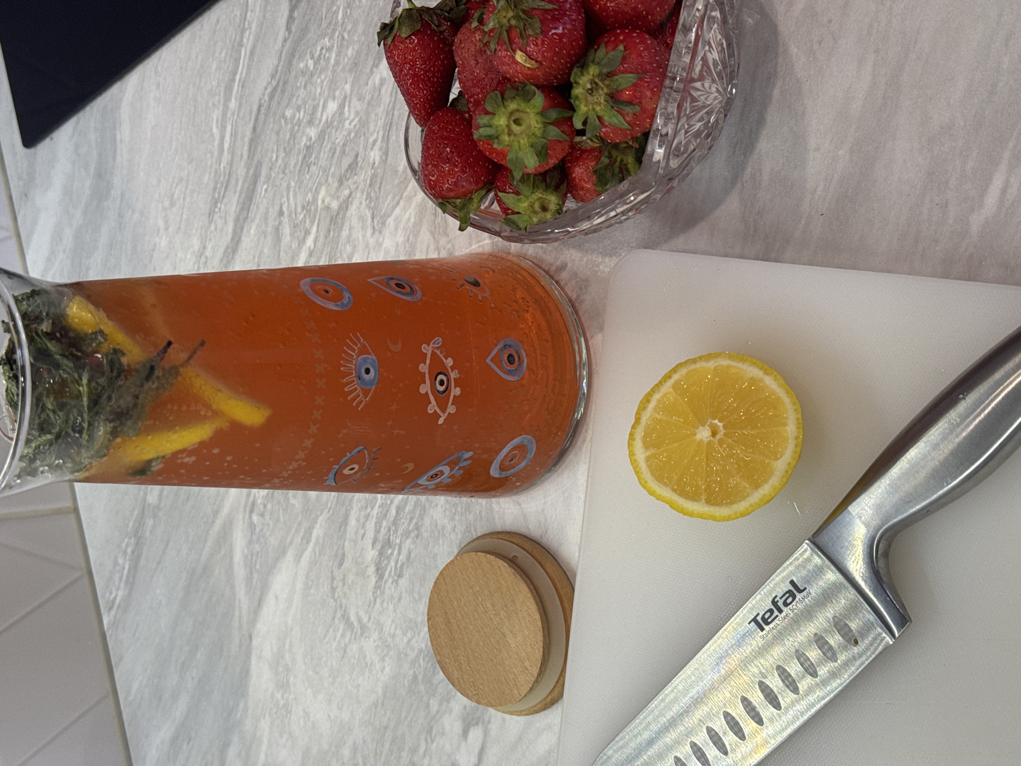 On a gray marble countertop, a clear glass pitcher filled with orange liquid containing herbs and lemon slices is oriented horizontally. Beside the pitcher is a glass bowl filled with strawberries. To the right, a halved lemon lies on a white cutting board, next to a silver Tefal knife. Also, there are some wooden coasters. The location is not discernible.