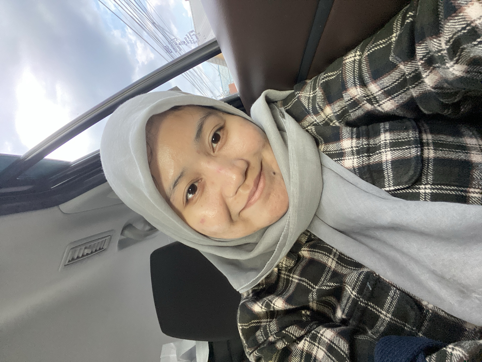 A woman wearing a light gray hijab and a black and white plaid shirt is inside a vehicle. She is looking at the camera and smiling. The vehicle appears to be an SUV, with dark brown leather seats visible. The background shows the sky with clouds and power lines, suggesting an outdoor setting within a populated area. The image was taken in Banjarbaru, Indonesia.