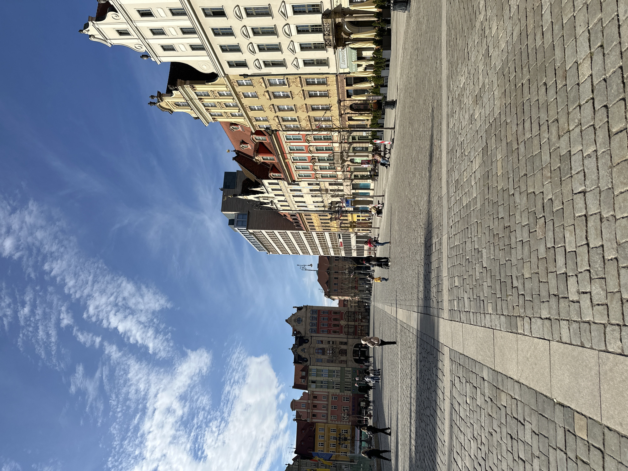 The image depicts a street scene in Wrocław, Poland. The perspective suggests the photo was taken at ground level looking lengthwise down a pedestrian square or street. Buildings with varying architectural styles line the left side of the frame, ranging from ornate, colorful structures with intricate window details to taller, more modern buildings. People are walking along the paved street. The sky is bright blue with wispy clouds. The paving is gray and appears to be made of cobblestones or a similar material. The photo seems to be taken in daylight with strong sunlight.