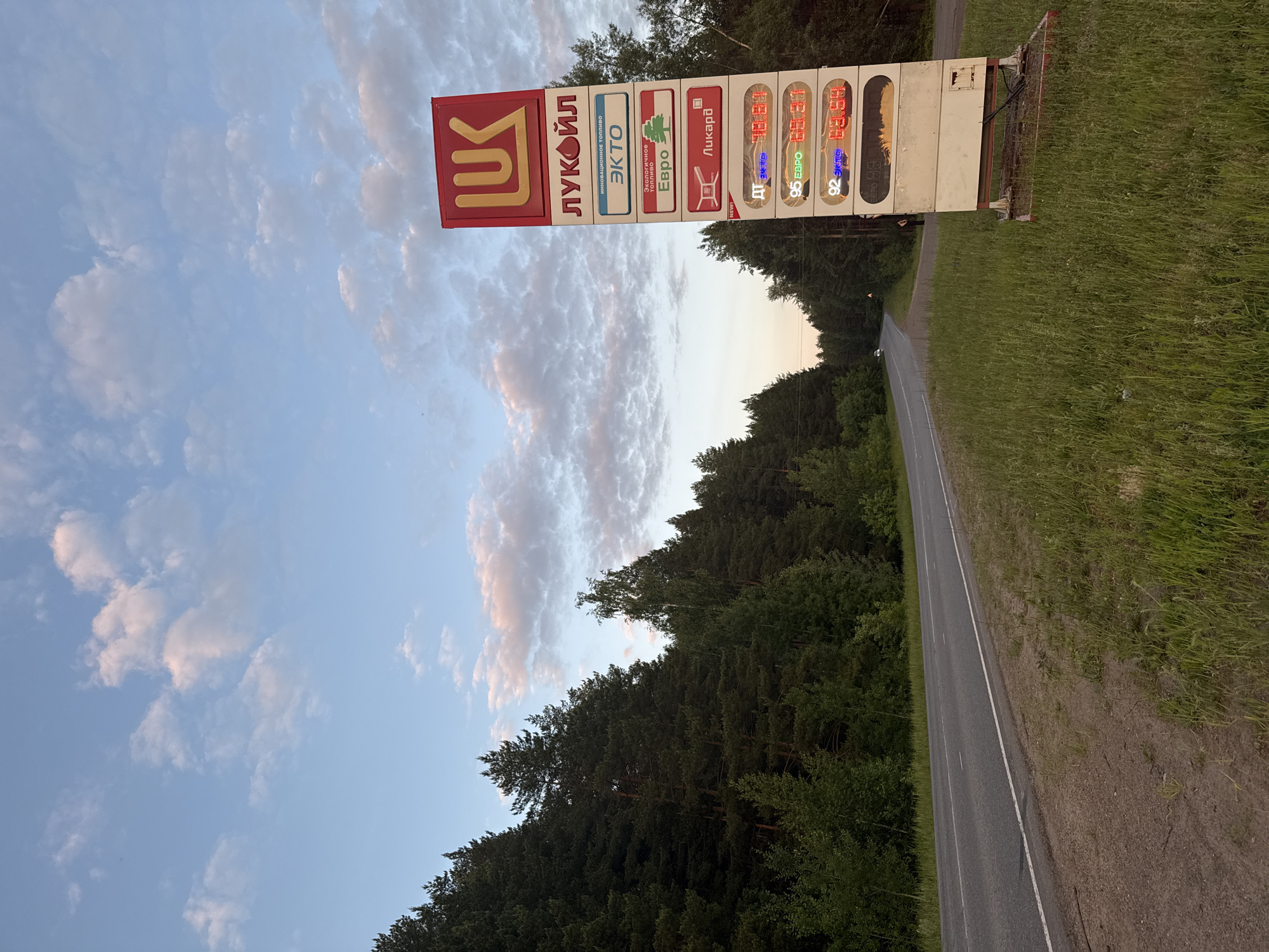 An image captures a roadside scene featuring a gas station price sign under a soft, cloudy sky. The sign belongs to LUKOIL (ЛУКОЙЛ), a prominent Russian oil company, confirming the general location of Камское, Russia.
The central subject is a tall, rectangular gas price board positioned beside an asphalt road that curves gently into the distance, flanked by a dense forest of coniferous trees on one side and a grassy shoulder on the other. The sky is a light blue with scattered, wispy clouds displaying hints of pink and orange, suggesting a late afternoon or early evening setting.
The LUKOIL sign is predominantly red and white. The top section features the LUKOIL logo. Below it, various fuel types and branding messages are displayed vertically. Visible text includes:
- "ЛУКОЙЛ" (LUKOIL)
- "ИННОВАЦИОННОЕ ТОПЛИВО" (Innovative Fuel)
- "ЭКТО" (ECTO)
- "ДОЛГОЛЕТИЕ МОТОРУ" (Longevity for the Engine)
- "Евро" (Euro)
- "ЛИКАРД" (LICARD)
- "NEW!"
Digital displays on the right side of the sign show fuel prices:
- "ДТ ЭКТО" (Diesel ECTO): 48.88
- "95 ЕВРО" (95 Euro): 50.94
- "92 ЭКТО" (92 ECTO): 46.84
- The lowest display is partially visible, showing "ЕВРО" and what appears to be "100" with prices partially cut off, but possibly starting with "57".
No people or vehicles are clearly visible, but the presence of the sign and road implies an area of transit. The overall atmosphere is tranquil, with natural elements framing the commercial sign.
