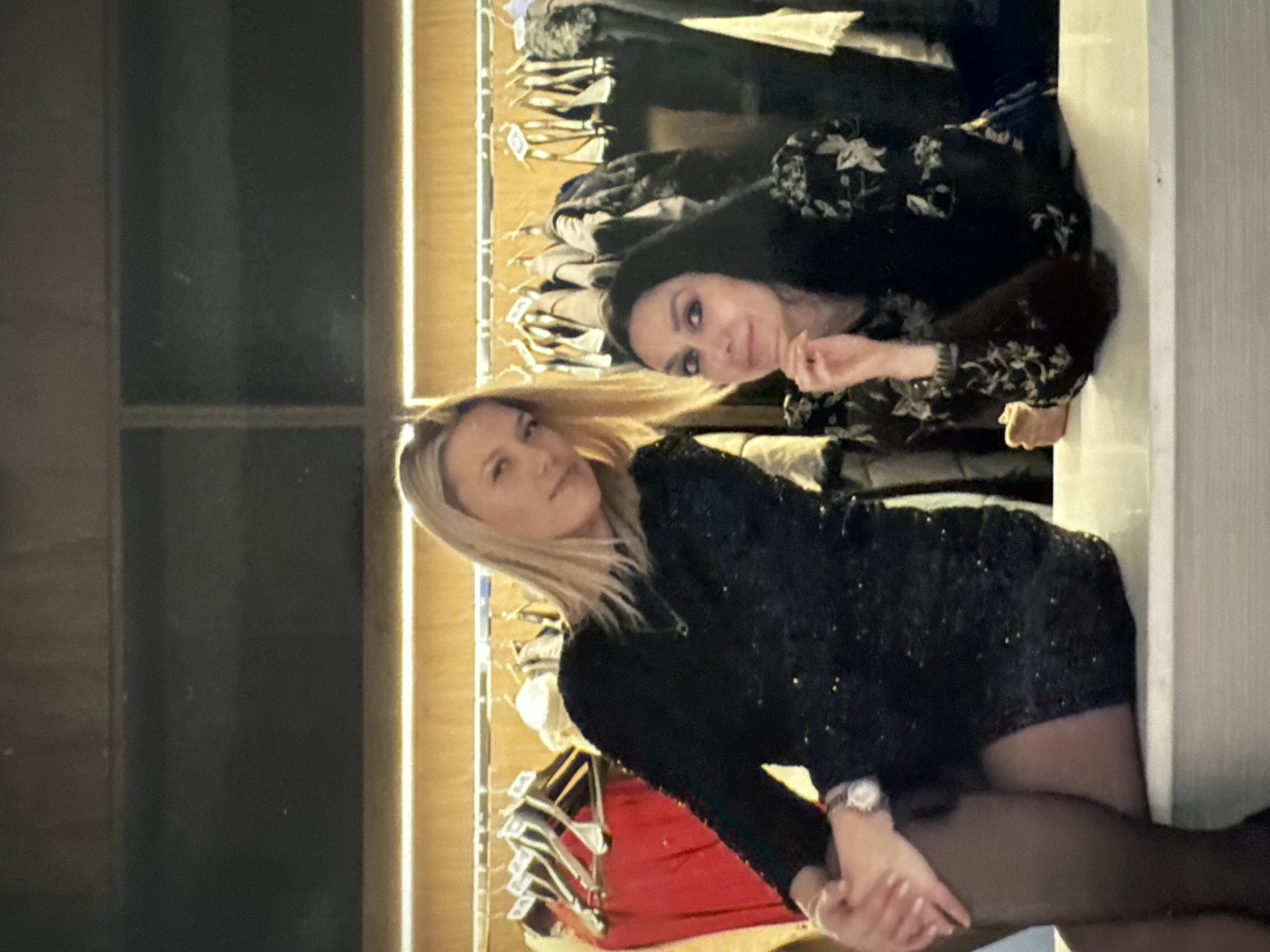 Two women are posed in front of a rack of clothing in what appears to be a boutique or a storage area. The woman on the left, with blonde hair, wears a black sequined dress and dark tights, and is seated with her legs crossed. She has a watch on her left wrist. The woman on the right, with dark hair, wears a black dress with a floral pattern and is leaning forward with her hand on her chin. The background shows a well-lit clothing rack with various garments, suggesting a commercial setting. The scene is captured indoors, with artificial lighting illuminating the area. This image was taken in Sofia, Bulgaria.