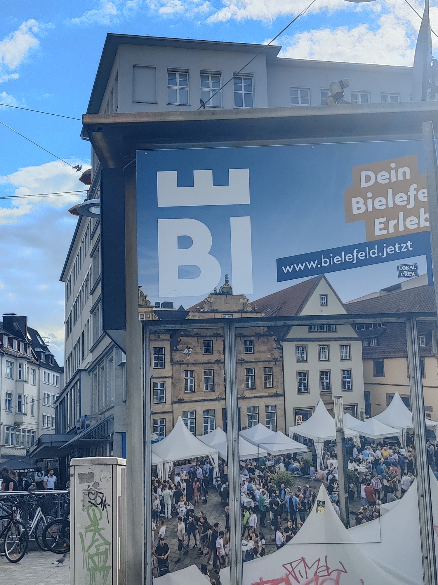 This image captures an outdoor scene in what appears to be a city square or street in Bielefeld, Germany. The sky is partly cloudy, suggesting it is daytime.
In the foreground, a large advertisement board dominates the view. The board displays a blue background with white text and graphics. A prominent white castle silhouette is visible, with the letters "BI" superimposed on it, likely representing "Bielefeld". To the right, the text "Dein Bielefe Erleb" is partially visible, suggesting a promotional campaign for experiencing Bielefeld. Below this, a blue banner contains the website address "www.bielefeld.jetzt" and a small logo with the text "LOKAL CREW". The lower portion of the advertisement features a reflection of a crowded outdoor event with many white tents.
The reflection reveals a bustling scene with numerous people gathered. They appear to be enjoying themselves, with some walking, some sitting at tables under the tents, and others socializing. The presence of tents suggests a market, festival, or outdoor fair.
In the background, to the left, a modern building with multiple floors and windows is visible. Further left, older, more traditional European-style buildings with pitched roofs can be seen. The scene is framed by overhead wires and a metal structure supporting the advertisement.
To the left of the advertisement board, a metal pole with some graffiti, primarily in green, is present. Several bicycles are parked near a sign that reads "STARBUCKS", indicating a commercial area.
The overall impression is a lively urban environment on a pleasant day, with a focus on promoting the city of Bielefeld and showcasing a community event.