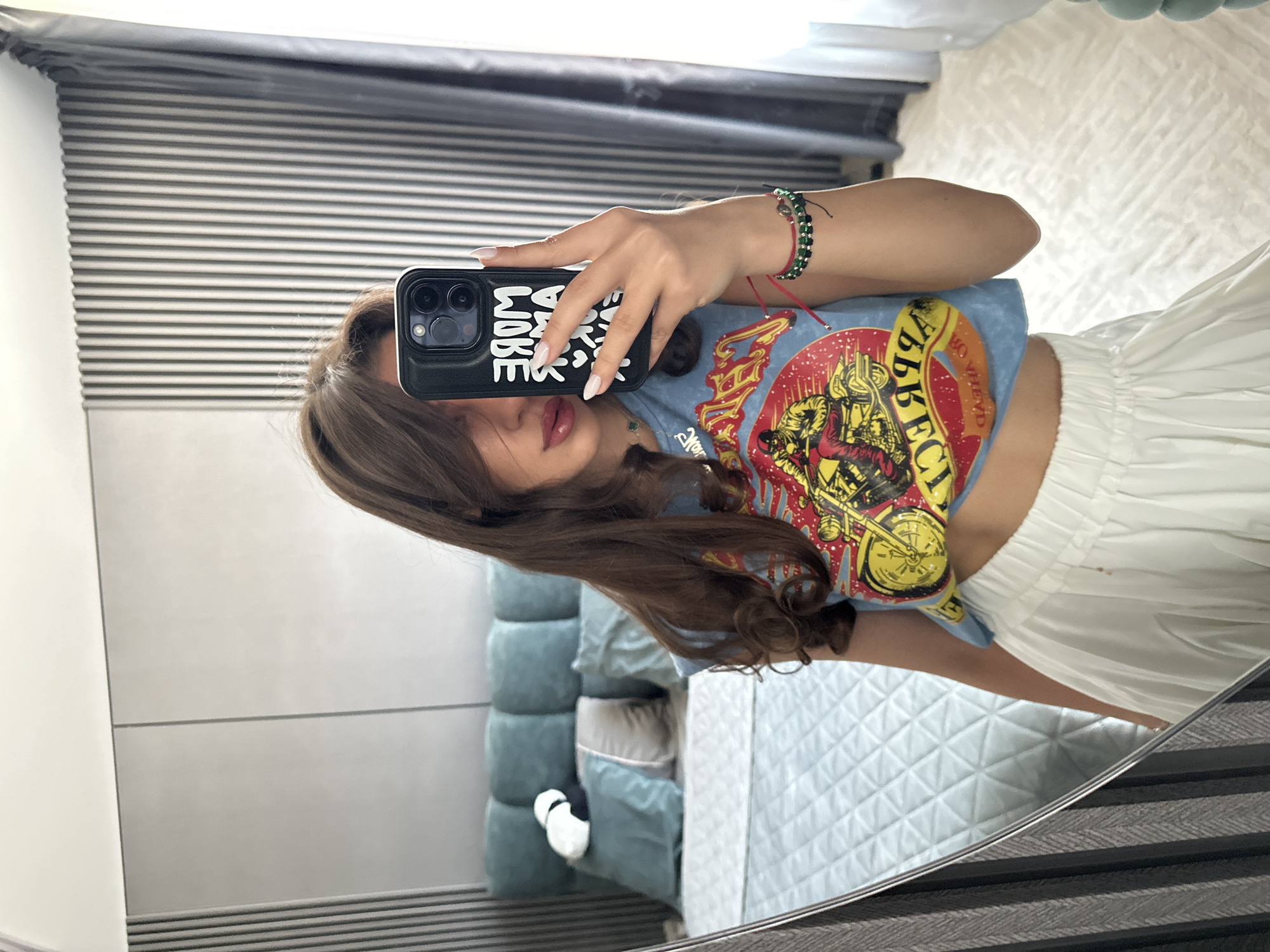 A young woman with long, brown hair, wearing a light blue cropped t-shirt with a motorcycle graphic and a white pleated skirt, takes a selfie in a bedroom. She is holding a black smartphone in her right hand, which is partially obscuring her face. Her left arm is visible, with a bracelet featuring green beads and a red string. The room features a bed with a light blue quilted comforter and a teal upholstered headboard with decorative pillows, one of which is a white and black panda. A large mirror is positioned to the left of the frame, reflecting the scene. The background includes a wall with vertical gray paneling and a gray curtain. The overall impression is that of a casual, personal moment captured in a modern bedroom environment. The location in Sofia, Bulgaria, is not discernible from the visual information provided.