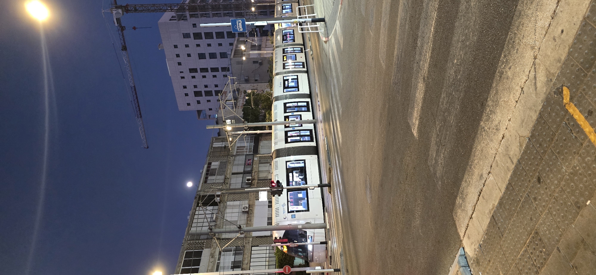 This image depicts a modern tram operating at night in Bat Yam, Israel. The tram, a white articulated vehicle with illuminated windows, is traveling along tracks on a city street. The street has a marked crosswalk in the foreground, indicating a pedestrian crossing.
The background reveals a cityscape with multi-story buildings, some appearing residential with numerous windows and balconies. Construction cranes are visible in the distance, suggesting ongoing development in the area. Streetlights and the tram's interior lighting illuminate the scene, casting reflections on the wet asphalt of the road. The sky is dark blue, indicating nighttime, and a faint circular halo is visible around the moon or a bright light source. A traffic light is positioned near the tram tracks, showing a red signal. A blue traffic sign with a tram symbol is also visible on a pole. The overall impression is of a functional urban environment at night, with public transportation playing a prominent role.