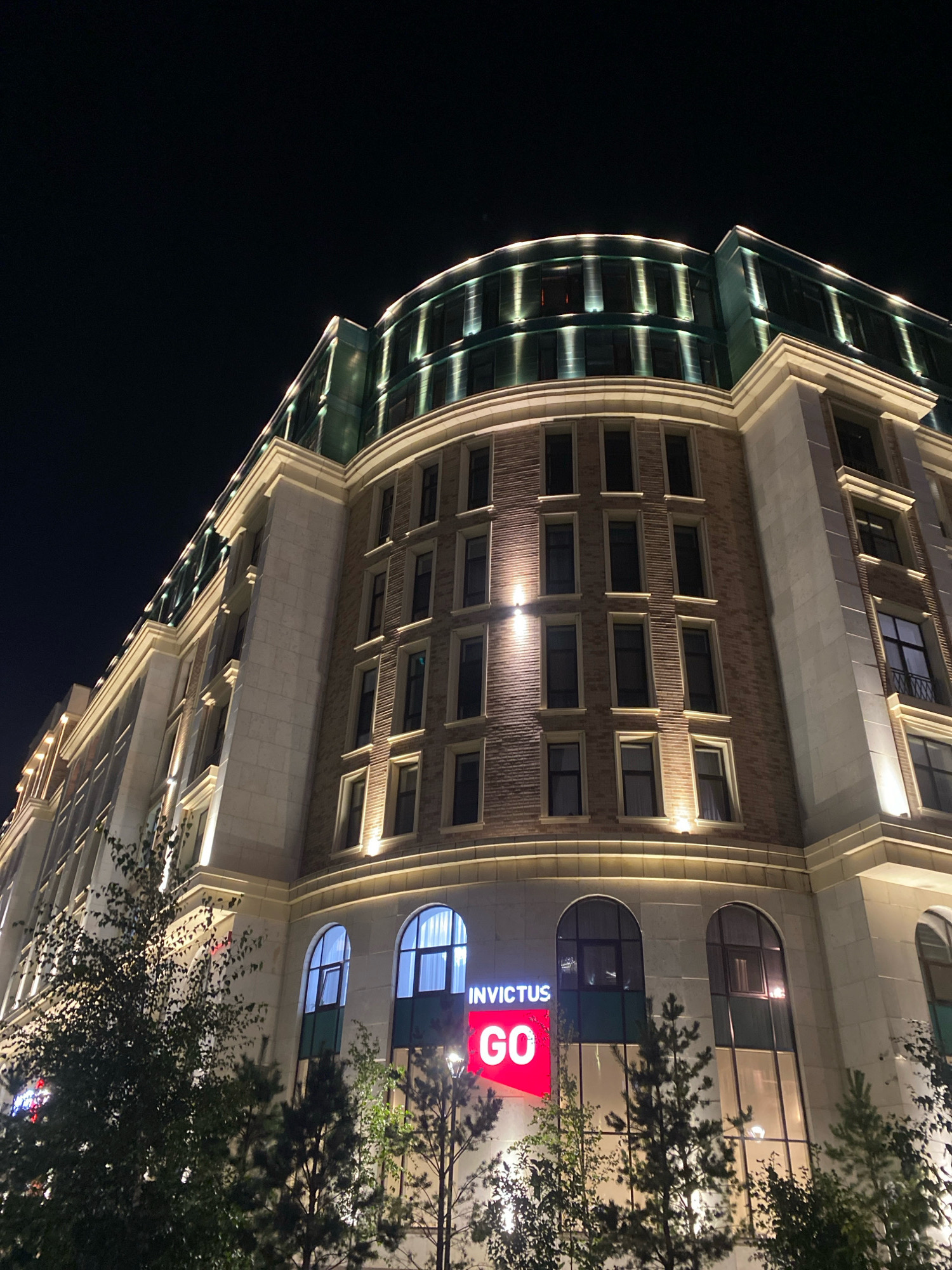 The image shows a modern, multi-story building at night, illuminated by external lighting. The building features a curved facade on the upper floors, with a green, reflective roofline accented by bright lights. The lower floors have a brick exterior with rows of windows. A prominent red sign displaying "INVICTUS GO" is visible on the lower part of the building. Trees and foliage are in the foreground, partially obscuring the lower levels. The sky is dark, indicating nighttime. This building is located in Astana, Kazakhstan.