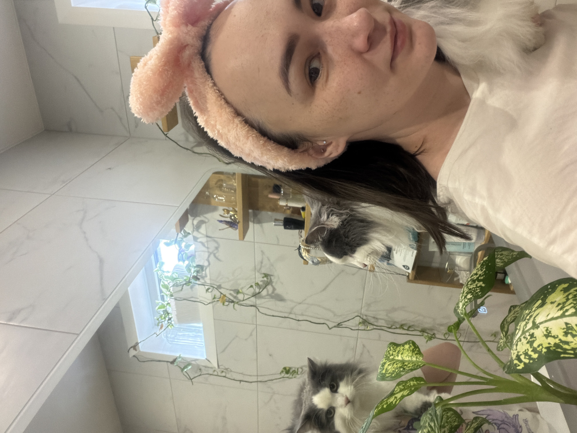 A woman with a pink headband is holding up a cell phone to take a selfie in a bathroom. The woman has brown hair and a white shirt. The bathroom has white tiled walls and a shelf with toiletries. A cat is visible in the reflection of a mirror, near a green potted plant. A window can be seen behind the cat and to the left. The location of this image is unknown and there is no information to suggest that it is in the city of Chaikovsky, Russia.