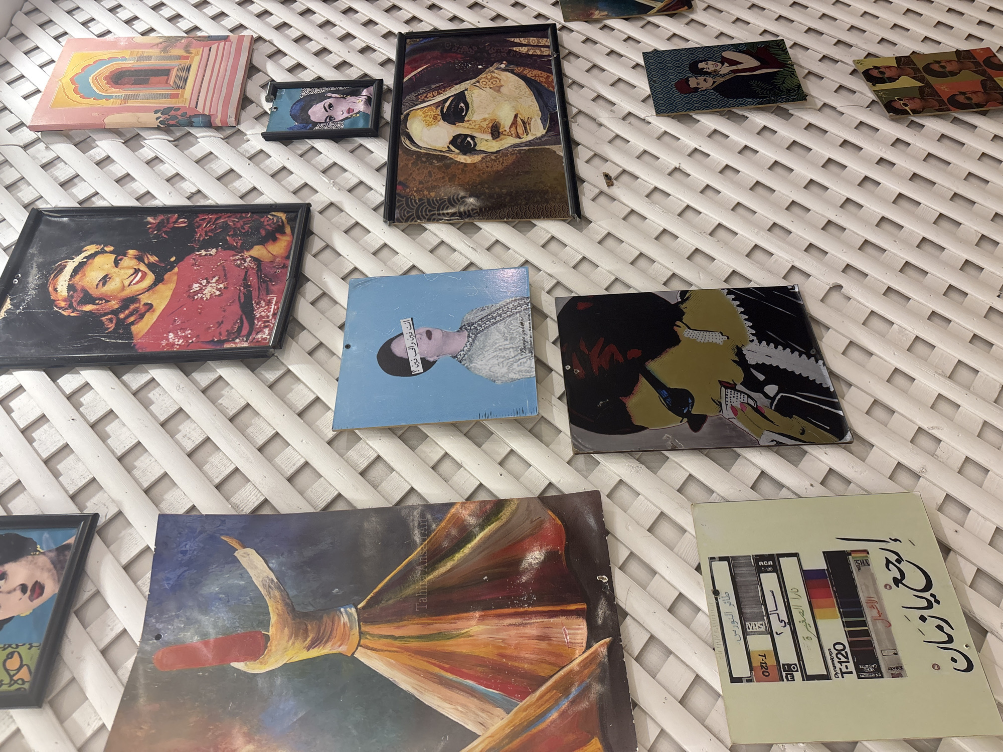 The image shows a wall with an assortment of framed artwork and photographs arranged in a somewhat haphazard manner against a white lattice background. The artwork includes a painting of a whirling dervish, portraits of women in various styles, and a piece resembling cassette tape labels with Arabic calligraphy. The overall impression is of a eclectic collection displayed in an indoor setting. There is no indication of the city of Amman, Jordan within the image itself.