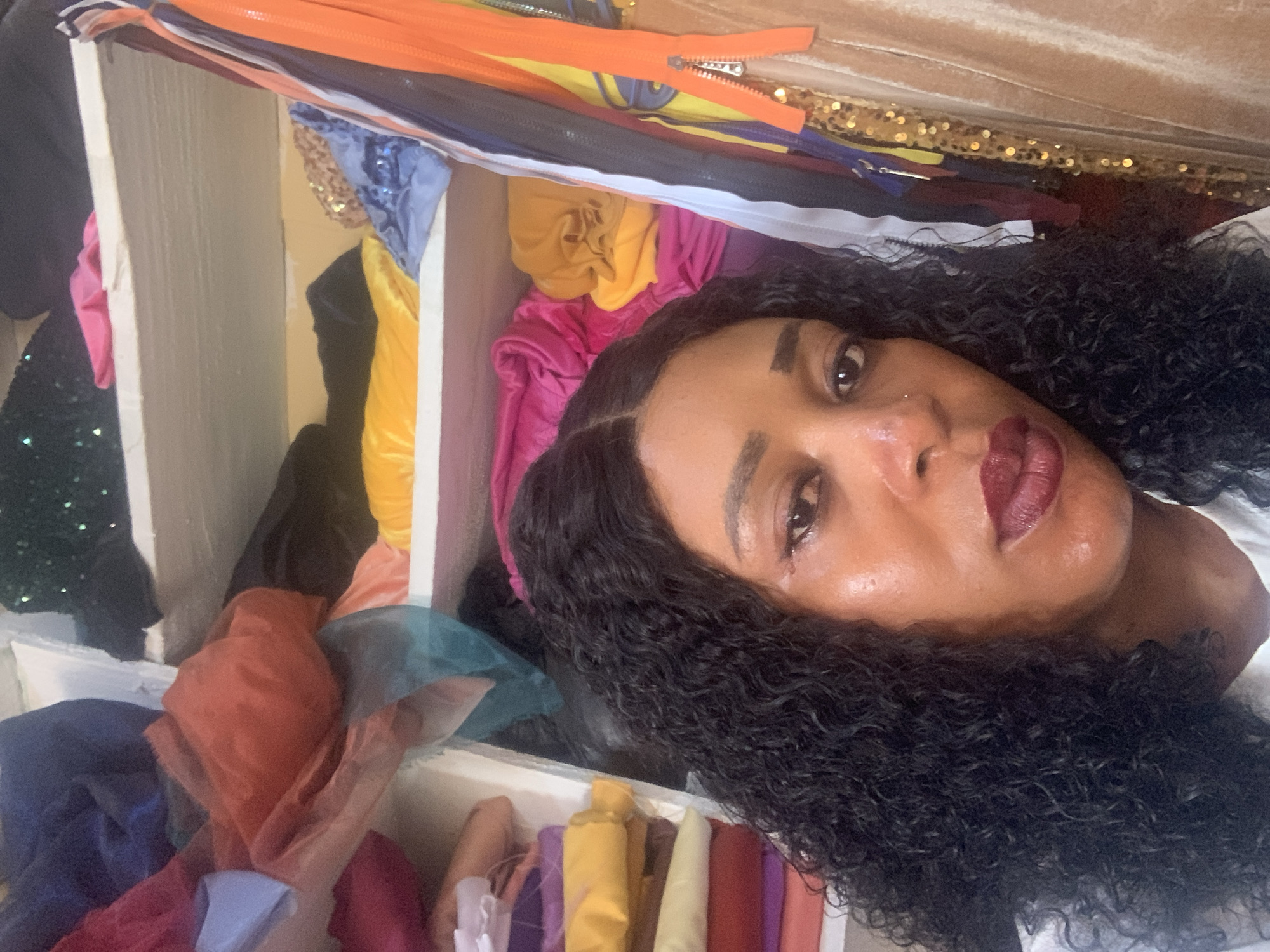The image shows a woman with curly dark hair and dark skin, wearing a white top, posing for a selfie in a room filled with fabrics and zippers. The room appears to be a storage space or workshop, possibly for sewing or fashion design, located in Sabon Tasha, Nigeria. Shelves are visible, stacked with various colorful fabrics in different textures and shades, including orange, yellow, blue, and pink. Zippers of various colors and sizes are also hanging or stored in the room, suggesting a focus on garment creation. The lighting in the room is bright, illuminating the woman's face and the colorful materials. The background shows a disorganized collection of textiles and sewing supplies, hinting at a creative and active environment.