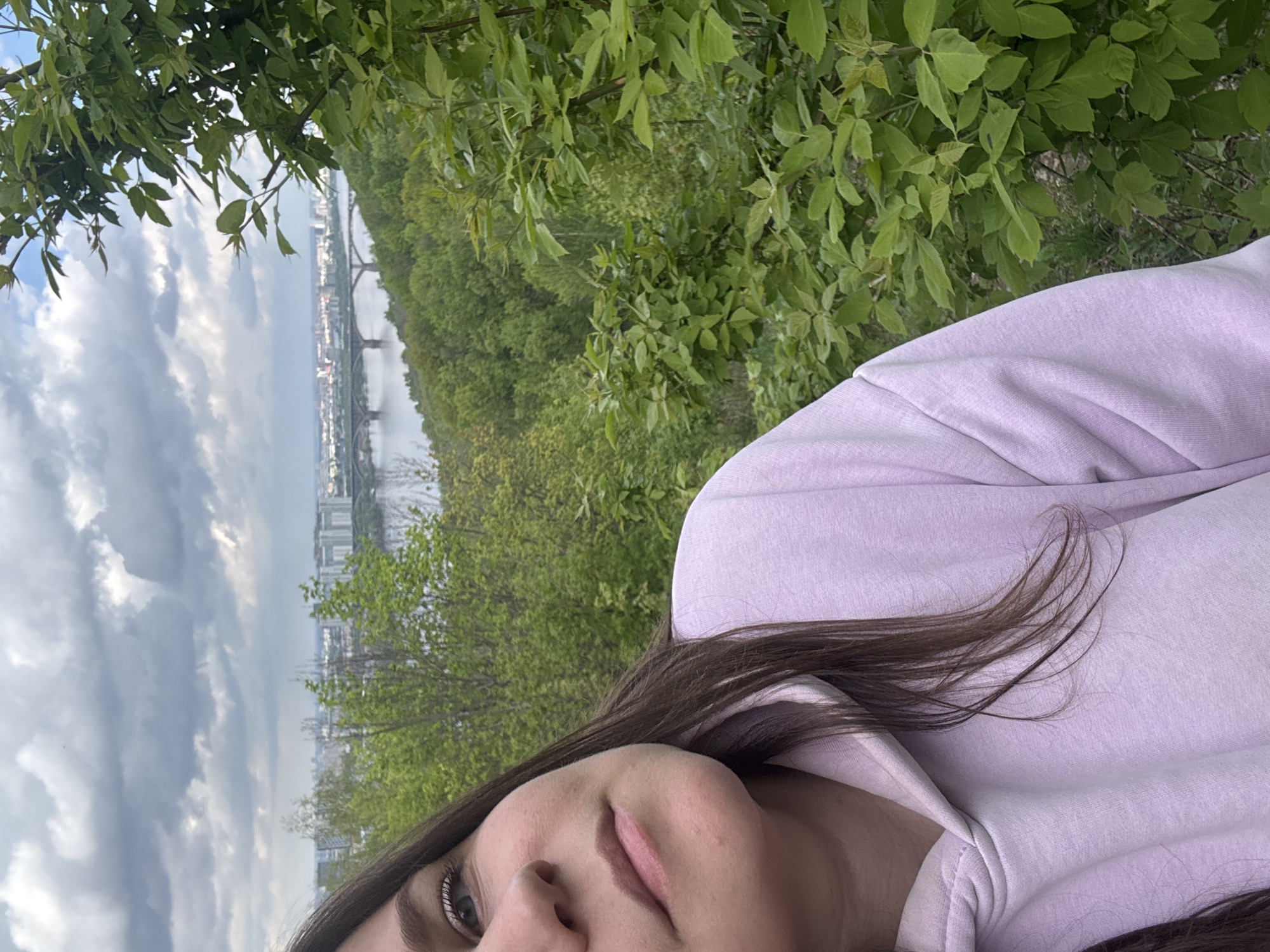 A selfie taken outdoors shows a person wearing a light purple hoodie with long brown hair. The background includes green trees, and in the distance, a river with a bridge and buildings can be seen under a cloudy sky. The location is identified as Nizhny Novgorod, Russia, due to the architectural features of the bridge and buildings.