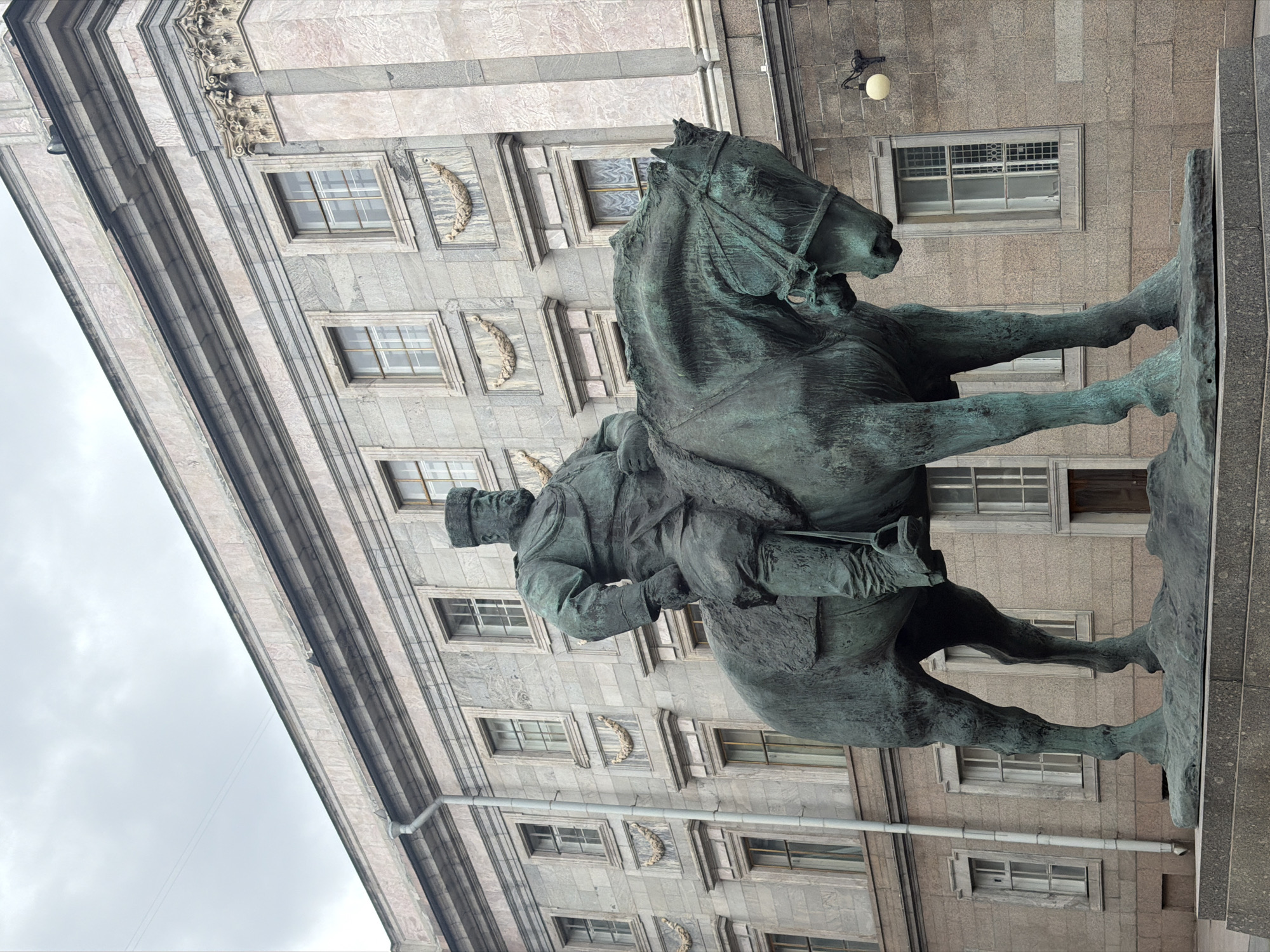An upright, eye-level photograph captures a majestic bronze equestrian statue against the backdrop of a grand, classical building under an overcast sky in Saint Petersburg, Russia. The statue depicts a distinguished military figure, likely a historical monarch, astride a horse. The rider is clad in a high-collared uniform, complete with a cap and a long coat, seated firmly in the saddle. The horse is rendered in a dynamic pose, with one front leg raised and its head slightly turned, conveying a sense of movement or readiness. The bronze material of the statue exhibits a characteristic green patina, indicative of its age. It stands atop a sturdy, dark stone pedestal.
The building behind the monument is an imposing structure of light-grey, textured stone, possibly granite or marble. Its facade is adorned with multiple rectangular windows, each framed by elaborate stone moldings. Between some windows, decorative relief carvings resembling laurel wreaths are visible. The architecture includes prominent cornices and structured pilasters, emphasizing its classical grandeur. The sky above is a uniform light grey, signifying an overcast daytime. A simple wall-mounted lamp is affixed to the building's facade. No visible text is present in the image. The scene evokes the monumental and historical character of Saint Petersburg.