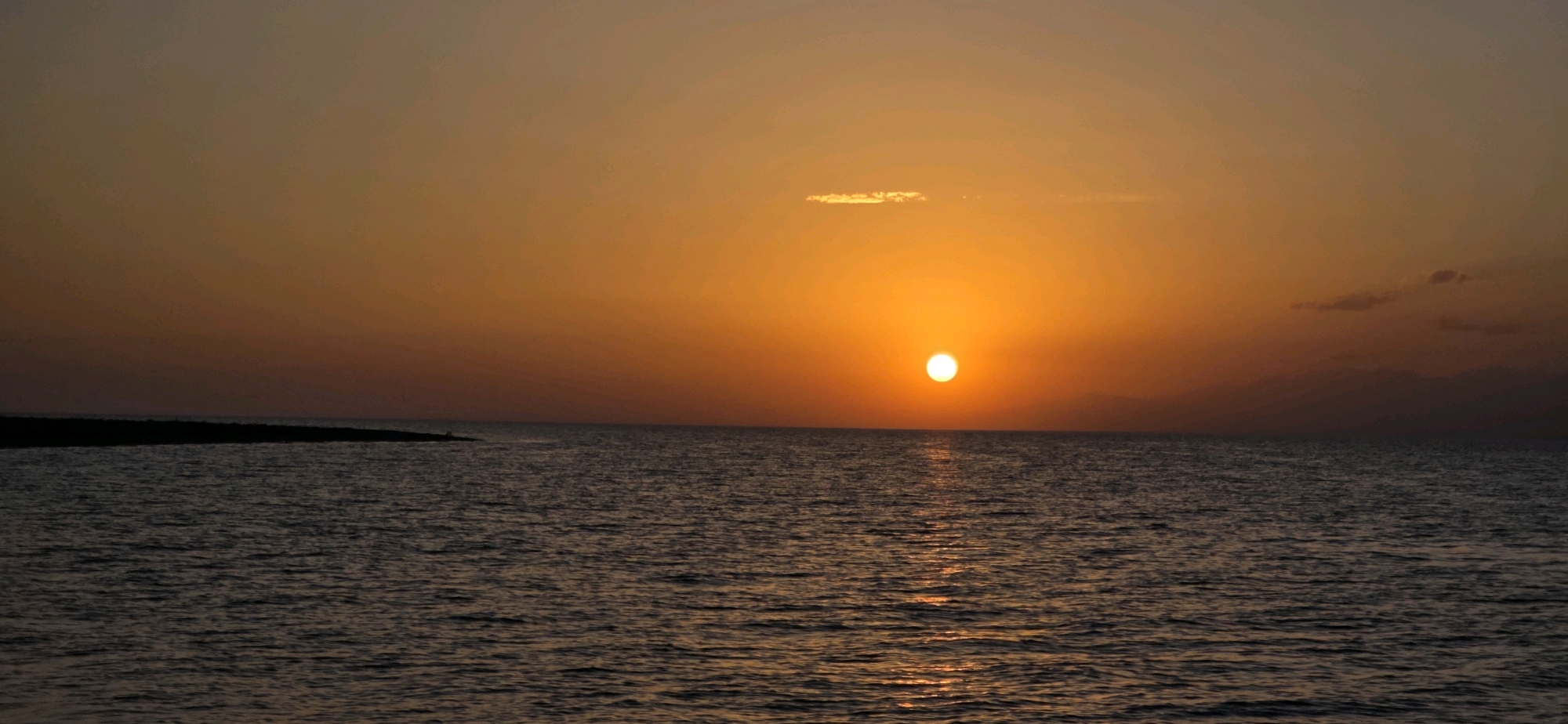 The image shows a sunset over a large body of water, likely a lake or sea. The sun is a bright orange orb, low on the horizon, casting a warm glow across the sky and reflecting on the rippling water. The sky transitions from a vibrant orange near the sun to a more muted brown and grey at the top of the frame. In the distance, a dark landmass is visible on the left side of the image, potentially a shoreline or island. The water is dark and appears calm with small waves. The overall impression is one of tranquility and natural beauty. There are no people, animals, or man-made structures clearly visible in the image. The scene is set in the city of Basbeshik, Kyrgyzstan.