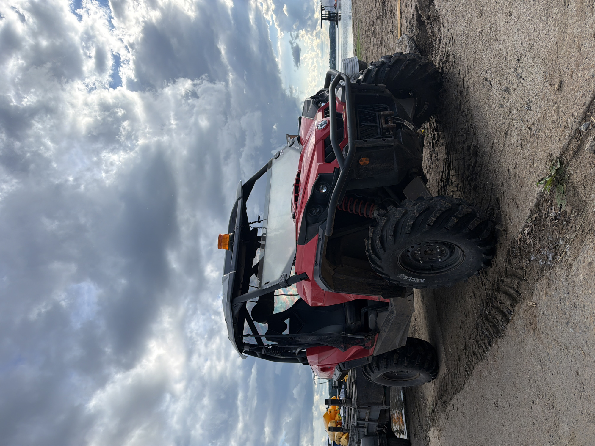 A red and black four-wheel all-terrain vehicle is parked on a sandy surface with tire tracks, possibly a beach or construction site. In the background is a body of water and a structure that could be a dock or pier. The sky is cloudy. The location is unknown, but based on the prompt, it could be near Votkinsk, Russia.