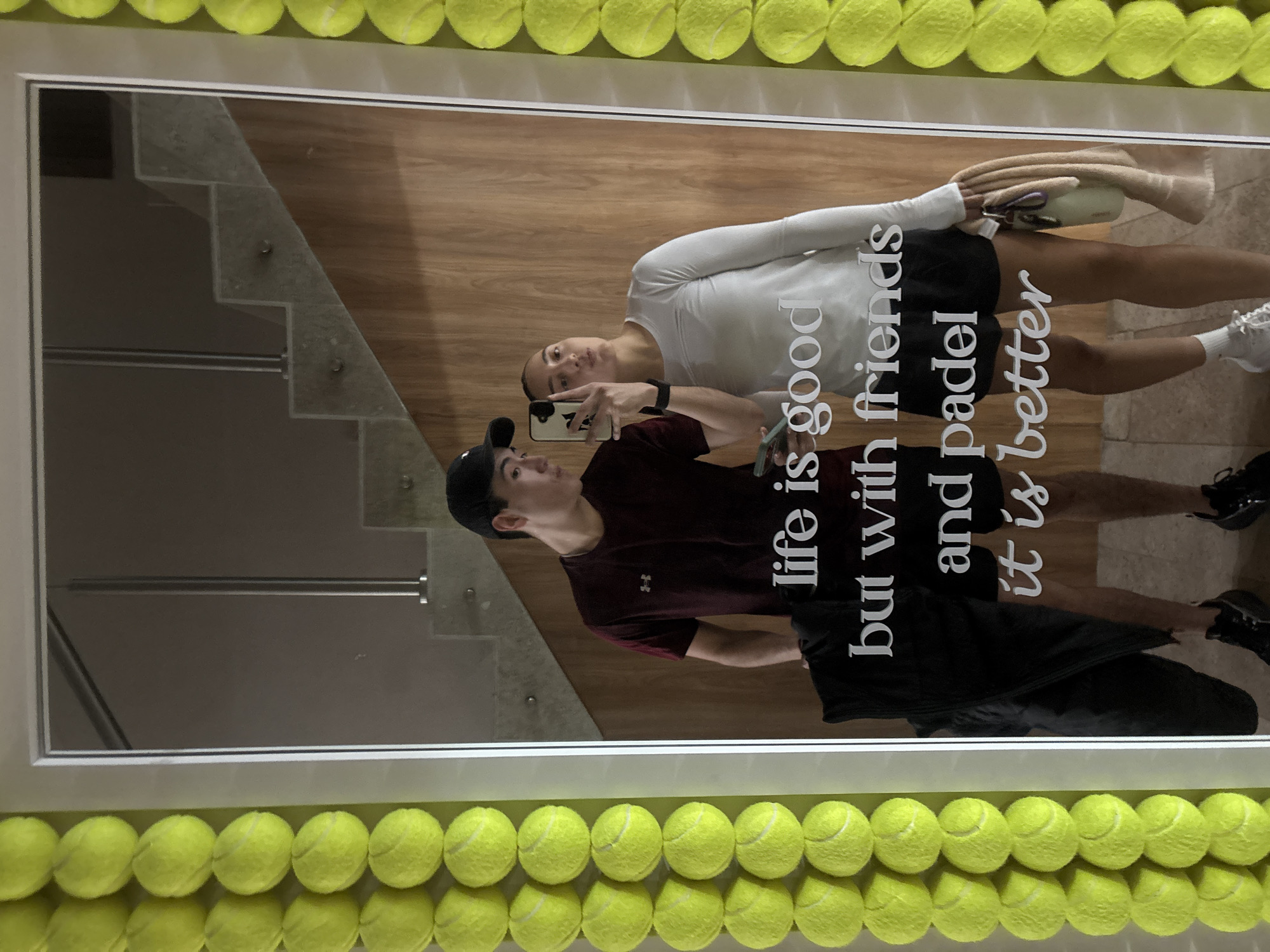 Two individuals are captured in a mirror selfie. A male figure, wearing a maroon shirt and a black baseball cap, is holding a smartphone and taking the photograph. A female figure, dressed in a white long-sleeved shirt and black shorts, is standing beside him. The reflection shows a stairwell and light wood paneling. The mirror is decorated with tennis balls around the frame and has the phrase "life is good but with friends and padel it is better" written on it. There is no indication the image was taken in San Bernardino Tlaxcalancingo, Mexico.