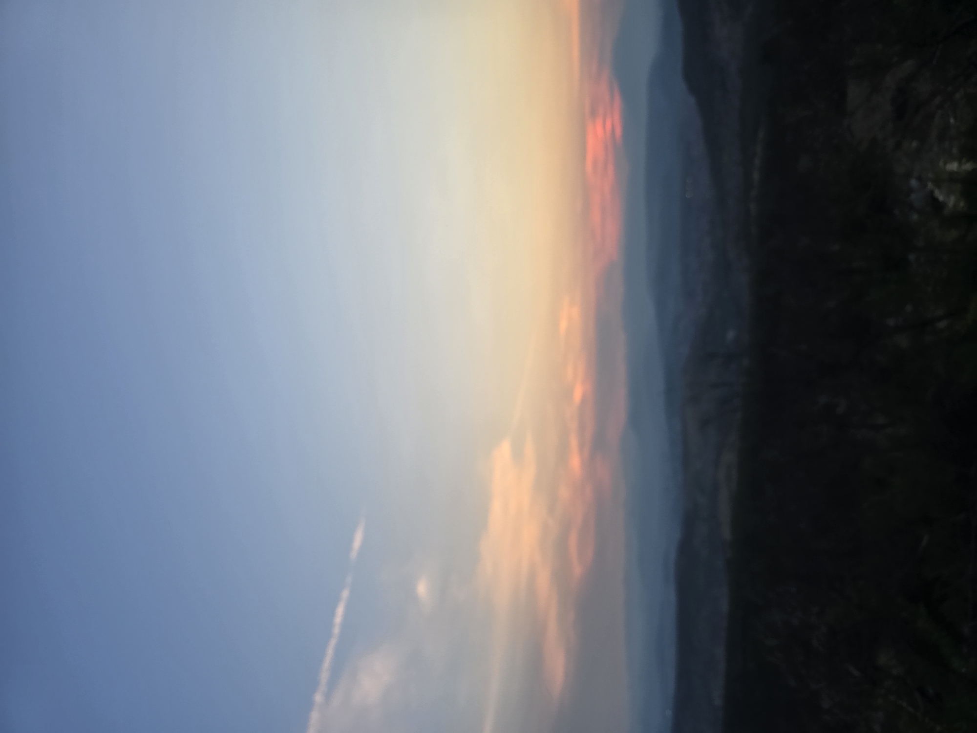 The image depicts a landscape at dusk, likely in or near Donja Vrbica, Montenegro, characterized by layered hills fading into the distance under a sky transitioning from blue to hues of orange and pink. In the foreground, the landscape is dark and appears to be covered in trees or dense vegetation. Beyond this, rolling hills are visible, with the furthest range appearing as faint blue silhouettes against the horizon. The sky exhibits a gradient, with a clear blue at the top, gradually warming to soft orange and pink tones closer to the horizon, suggesting the setting sun. A few wispy clouds are present in the upper left quadrant. There are no people, animals, or specific man-made structures clearly identifiable in the image due to the distance and lighting conditions. The overall impression is one of a serene, natural environment during twilight.