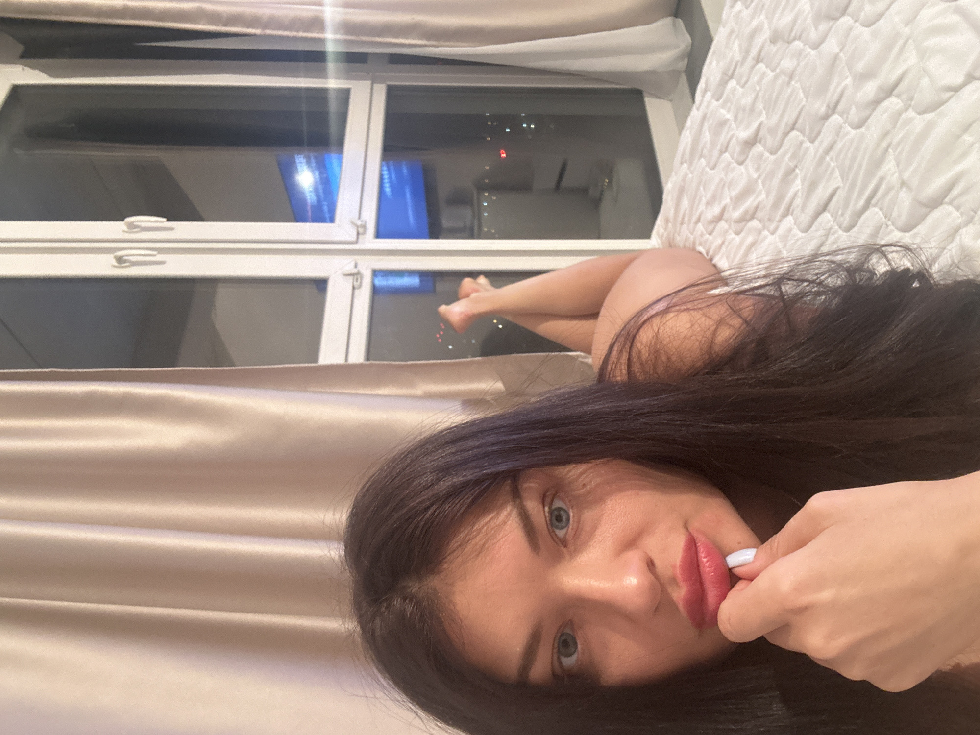 An upward-angled, close-up shot captures a young woman lying down in an indoor setting at night. She has long, dark hair, light blue eyes, and full, pink lips, looking directly at the viewer. Her right hand is positioned near her mouth, with a finger, possibly adorned with light-colored nail polish, gently touching her lips.
She is situated on a white, quilted mattress or duvet, which occupies the right side of the frame. In the background, her bare legs are visible, bent at the knees. Behind her, a large window with white frames reveals a dark nighttime cityscape, dotted with numerous lights from buildings, indicating an urban environment. A blue light source, possibly a television screen, reflects in the window glass. Light-toned (beige or tan) curtains frame the left side and top of the image, suggesting a bedroom or living area. The scene depicts a quiet evening within a high-rise building in what is known to be Krasnogorsk, Russia. No visible text is present.