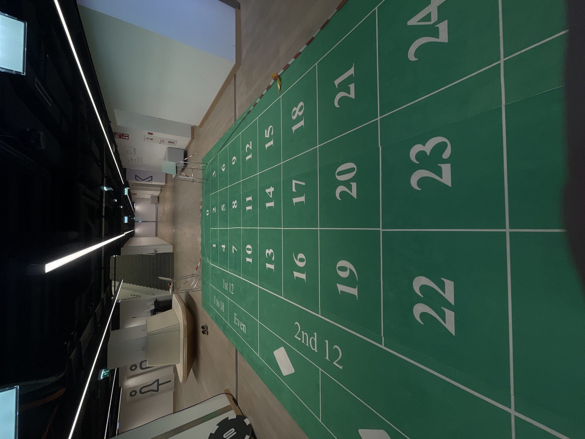 The image presents a tilted, high-angle view of a roulette betting layout, indoors in an unspecified location. The layout features a green surface divided into rectangular sections. The sections are labeled with white numerals and text indicating the numbers 1 through 24, as well as groupings such as "1st 12," "1 to 18," "Even," and "2nd 12." The overall orientation of the image is such that the roulette layout appears to extend away from the viewer. The layout is indoors in an urban area in Unknown, Unknown. The image includes modern interior architectural details such as ceiling-mounted light fixtures and wall signage.