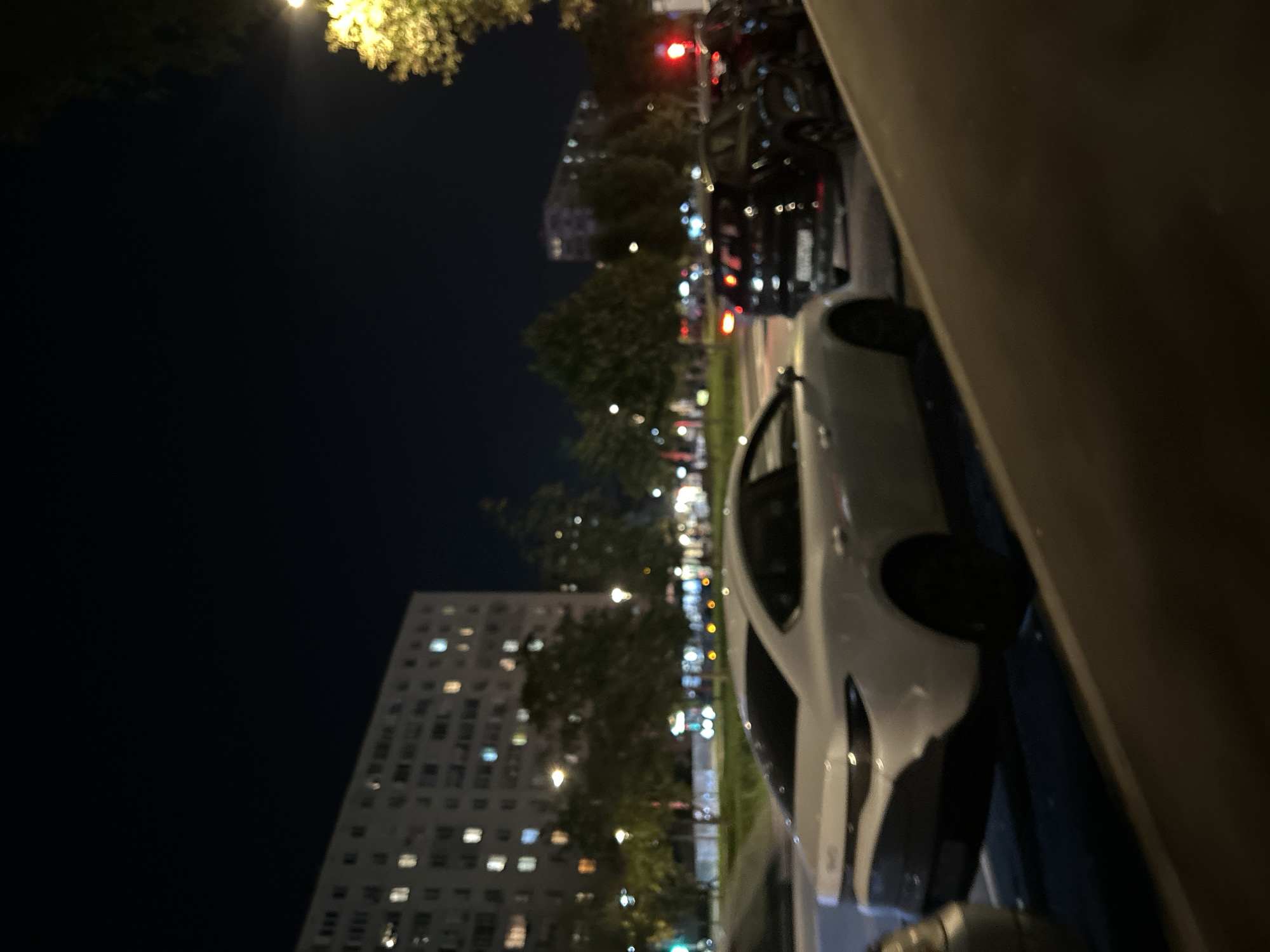 The image depicts a street scene at night, likely in a residential area. A white sedan is parked on the left side of the road, facing away from the viewer. Behind it, several other cars are parked, including a dark-colored SUV. In the background, a large apartment building with illuminated windows is visible, suggesting a densely populated urban environment. Trees line the street, and streetlights cast a yellow glow. The overall impression is one of a typical city night, with cars parked along the street and lights from buildings illuminating the darkness. The absence of any people or specific landmarks makes it difficult to pinpoint the exact location, but the architectural style of the apartment building and the presence of trees suggest a European city, possibly within Moscow, Russia.
