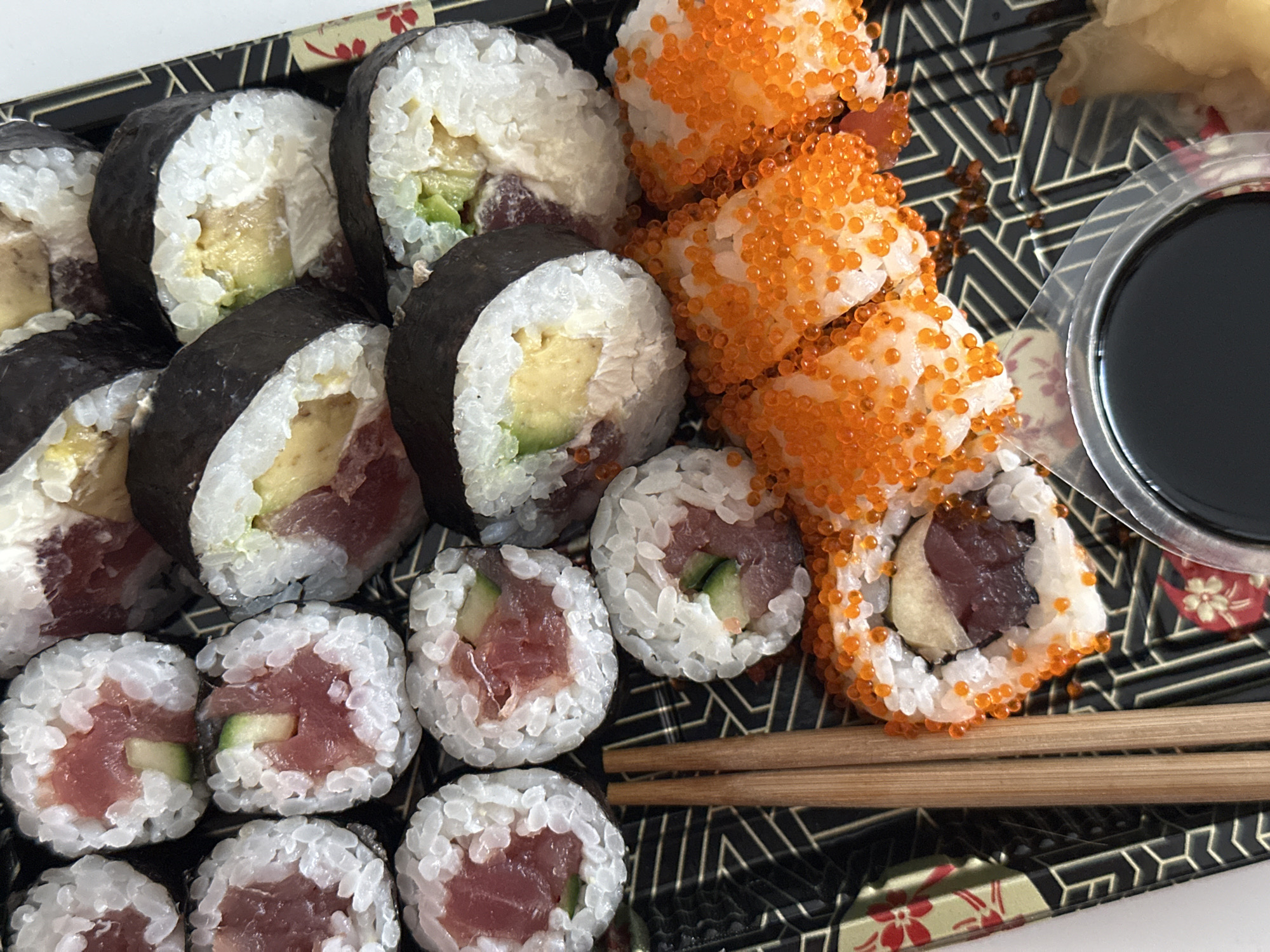 The image shows a tray of assorted sushi rolls. From left to right, there are rolls with nori seaweed, rice, tuna, avocado, and egg. To the right are rolls coated in orange fish roe. A small plastic container filled with soy sauce is also visible, along with chopsticks. The image does not contain information to verify the location.