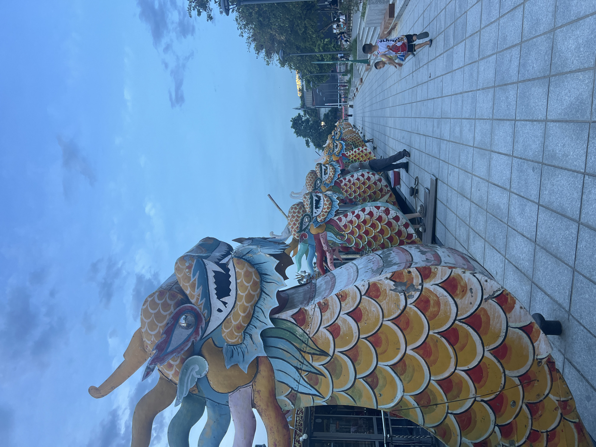 The image depicts a promenade along a waterfront, likely in the city of Thuận Hóa Ward, Vietnam, characterized by a row of large, colorful dragon boat heads. The dragon heads are intricately decorated with scales in shades of yellow, orange, red, and blue, featuring sharp teeth and elaborate horns. They are positioned in a line, facing towards the left of the frame, with their ornate tails extending out of view. The promenade is paved with light-colored square tiles and is lined with trees and lampposts on the right side. In the background, several people are visible, some seated on benches or standing near buildings. Two young boys are in the foreground on the right, one of whom is running. The sky is a hazy blue with scattered clouds, indicating it may be late afternoon or early evening.