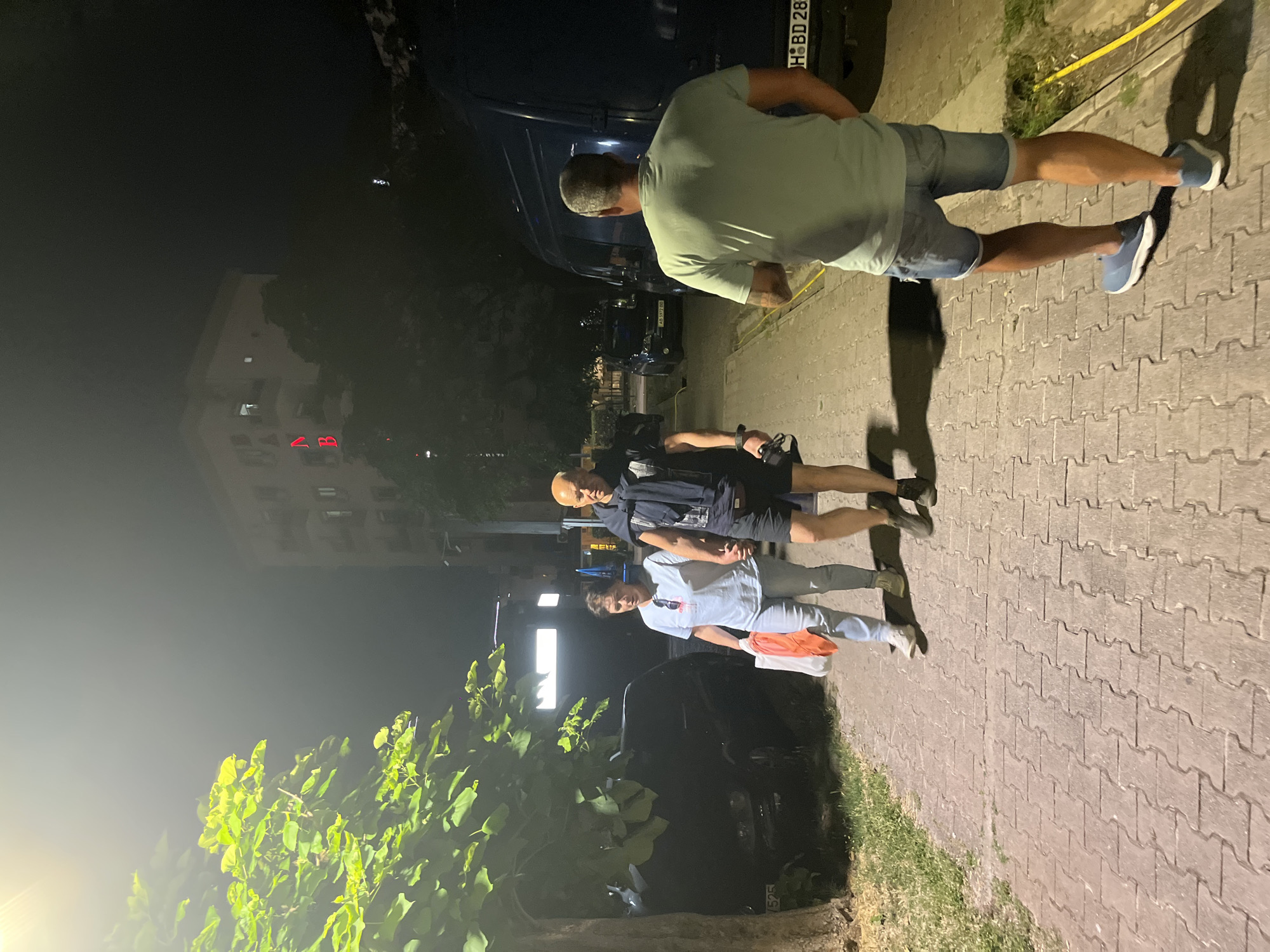 Three individuals are walking on a paved sidewalk at night. On the left, a woman and a man are walking hand-in-hand. The woman is wearing a light blue t-shirt, light blue jeans, and white sneakers, and she is carrying a white plastic bag and an orange item. The man is wearing a dark t-shirt, dark shorts, and dark shoes, and he has a dark blue jacket draped over his shoulders. He is also holding a black object in his right hand. In the foreground and facing away from the camera, a man is walking on the sidewalk. He is wearing a light green t-shirt and denim shorts, and blue sneakers. He is casting a long shadow on the ground. The sidewalk is lined with grass on the left and right. Several parked vehicles are visible in the background, including a dark SUV and a dark van. A multi-story building with illuminated windows is visible in the distance, with red neon lettering on its facade. The scene is illuminated by artificial lighting, casting shadows and creating a somewhat dimly lit atmosphere.
