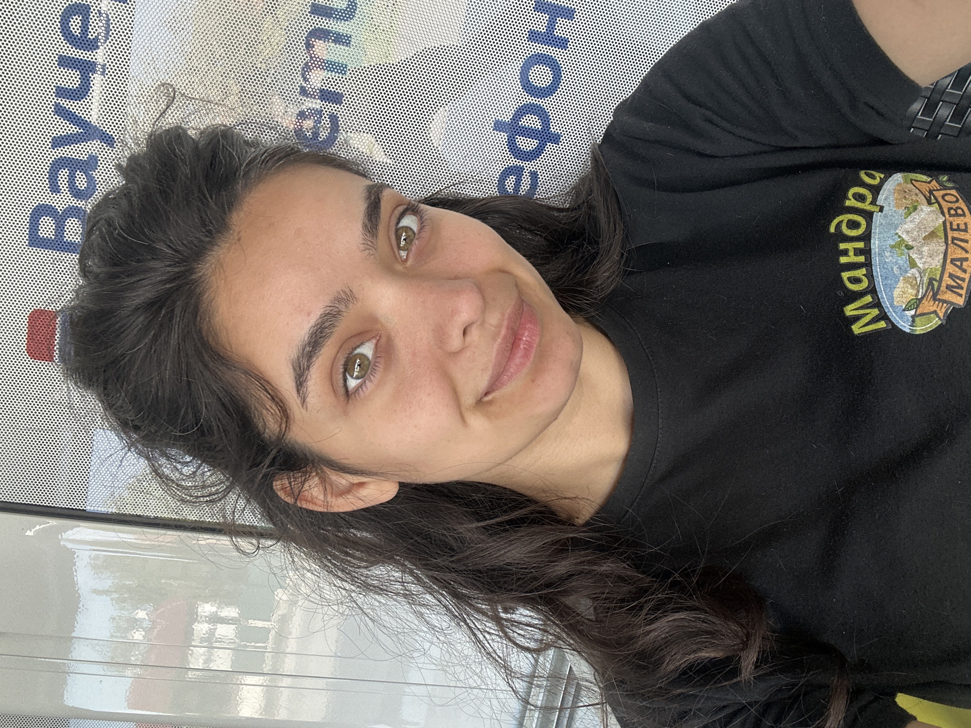 A young woman with dark, wavy hair and brown eyes is taking a selfie. She is wearing a black t-shirt with a logo on it. The logo is oval-shaped with a banner across the bottom. Inside the oval, there are images of food. The banner at the bottom says "MALEVO". The background appears to be a window with text and images visible through it. The text is in Cyrillic script and appears to be an advertisement. The woman is smiling and looking directly at the camera. The overall scene suggests an outdoor setting, possibly a cafe or shop in Malevo, Bulgaria, given the text on the t-shirt.