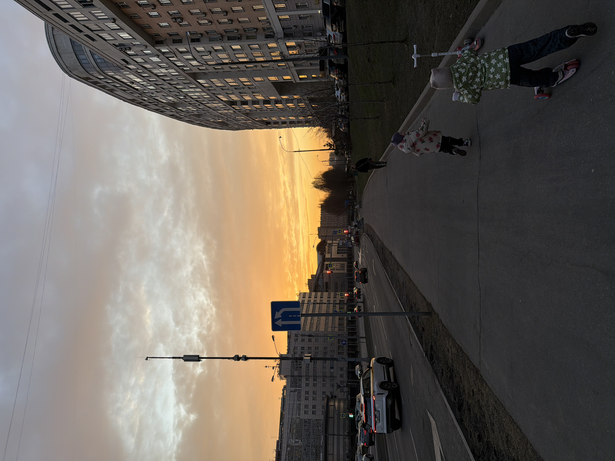 The image depicts an urban street scene in Khimki, Russia, during sunset or dusk. The sky displays a vibrant orange and yellow glow along the horizon, transitioning to a lighter grey with scattered clouds overhead.
In the foreground, a dark asphalt sidewalk extends from the lower right towards the center-left. Two young children are visible on the sidewalk, moving away from the camera. One child, wearing a light-colored, patterned jacket, is running. The second child, in a green floral jacket and light beanie, is riding a pink and black three-wheeled scooter. A white three-wheeled scooter is positioned stationary to the right of the running child. Further along the sidewalk, a single adult figure in dark clothing walks away from the viewer.
To the immediate right of the sidewalk is a narrow grassy strip with sparse, bare trees, bordering a large, multi-story, curved building. This building features numerous windows, some reflecting light from the sunset, and multiple external air conditioning units on its facade. Several parked cars are visible along its base.
To the left of the sidewalk is a multi-lane asphalt road with active vehicular traffic. A white crossover SUV, featuring yellow side accents and Russian license plate "728T 750", is prominent in the leftmost visible lane, traveling away from the camera. Other vehicles are present in various lanes, some displaying illuminated headlights. Street infrastructure includes a tall utility pole equipped with communications devices, and a blue rectangular road sign displaying two upward arrows, positioned between the sidewalk and the road. Red traffic lights are visible in the far distance. Additional multi-story urban buildings are discernible in the background, partially silhouetted against the sunset.