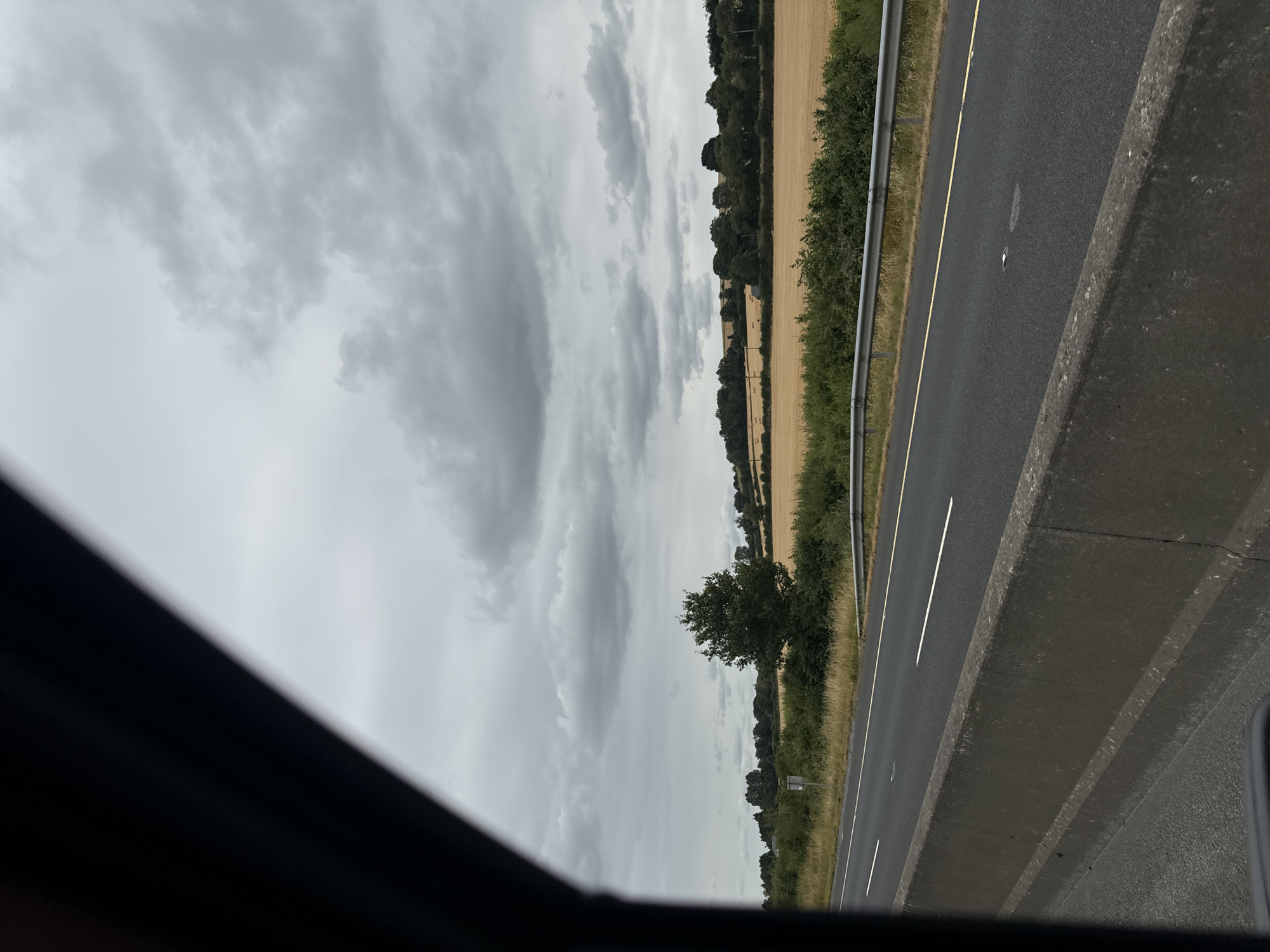 The image captures a view from a vehicle traveling on a highway, likely near Dunlavin, Ireland, given the agricultural landscape. The foreground is dominated by a concrete barrier and the asphalt of the highway with white lane markings. To the right of the road, a metal guardrail separates the highway from a grassy verge. Beyond the verge, a field of golden stubble, indicative of harvested crops, stretches across the landscape. A line of trees and bushes borders the field, with a solitary, mature tree standing prominently in the mid-ground. In the distance, rolling hills are visible, also lined with trees. The sky is overcast with heavy, gray clouds, suggesting a typically cloudy Irish day. There are no people or discernible actions taking place in the image; it is a still, environmental shot focused on the roadside and agricultural scenery.