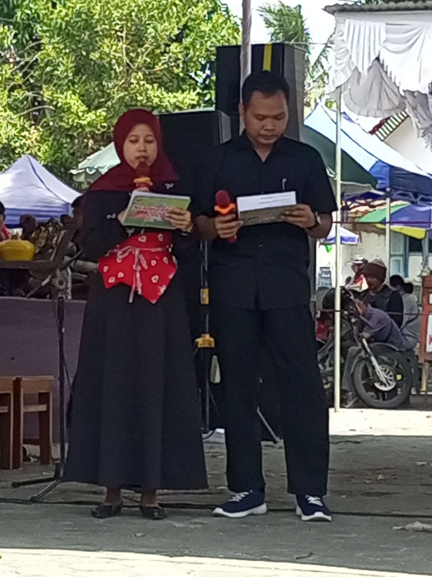 In Nanggulan, Indonesia, a woman wearing a red hijab and a long dark dress, accented with a red and white patterned sash, stands holding a microphone and a book. Beside her, a man in a black collared shirt and dark pants also holds a microphone and a document. They appear to be addressing an audience or participating in an event. In the background, various tents, a motorcycle, and trees are visible, suggesting an outdoor gathering.