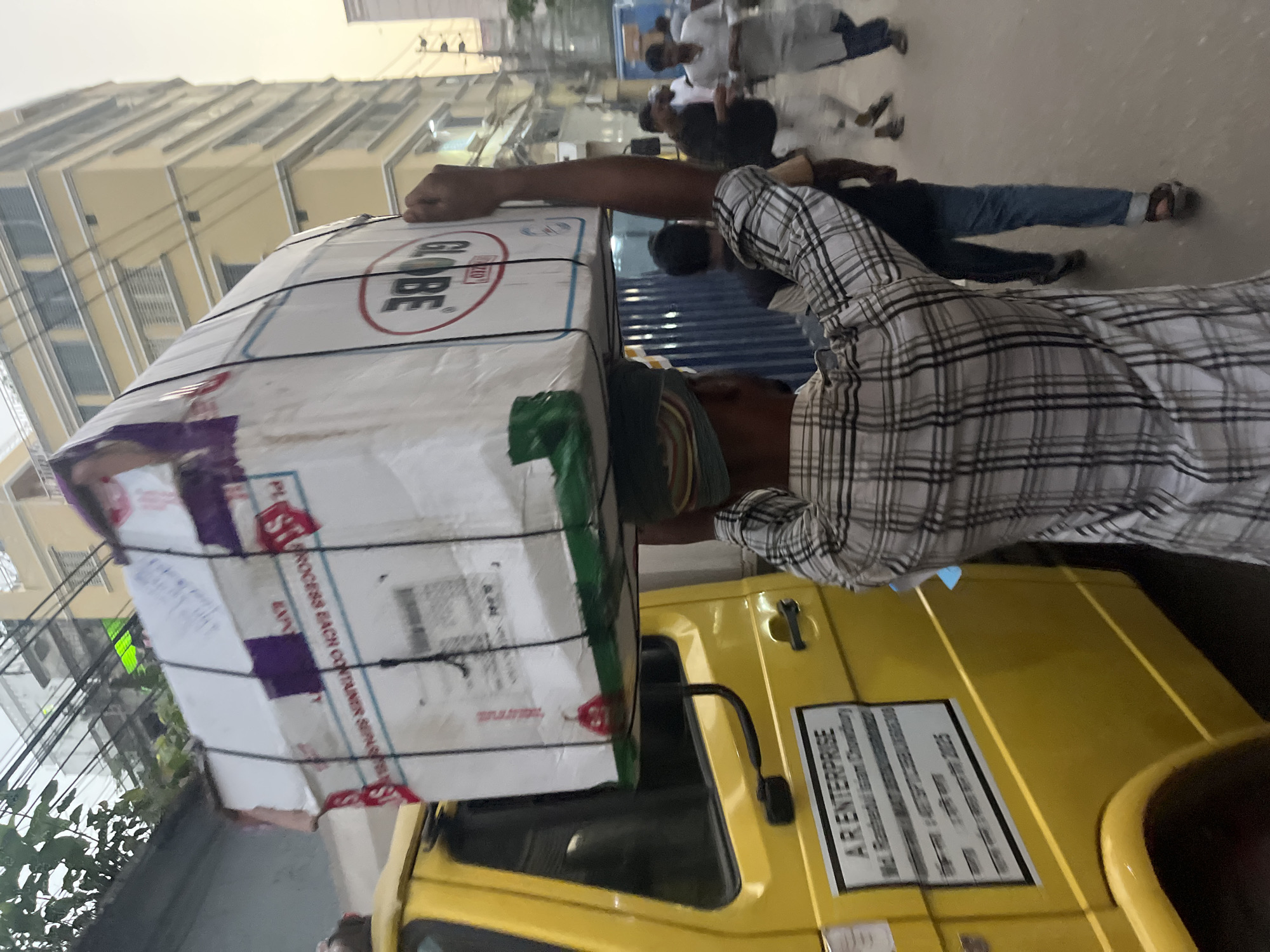 In Dhaka, Bangladesh, a man wearing a plaid shirt carries a large, white cardboard box labeled "GLOBE" on his head, which is secured with rope and padding. The box sits atop a yellow vehicle with "A R ENTERPRISE" printed on the side. Other people walk along the street in the background of the image. A tall building with multiple stories is visible as part of the background.