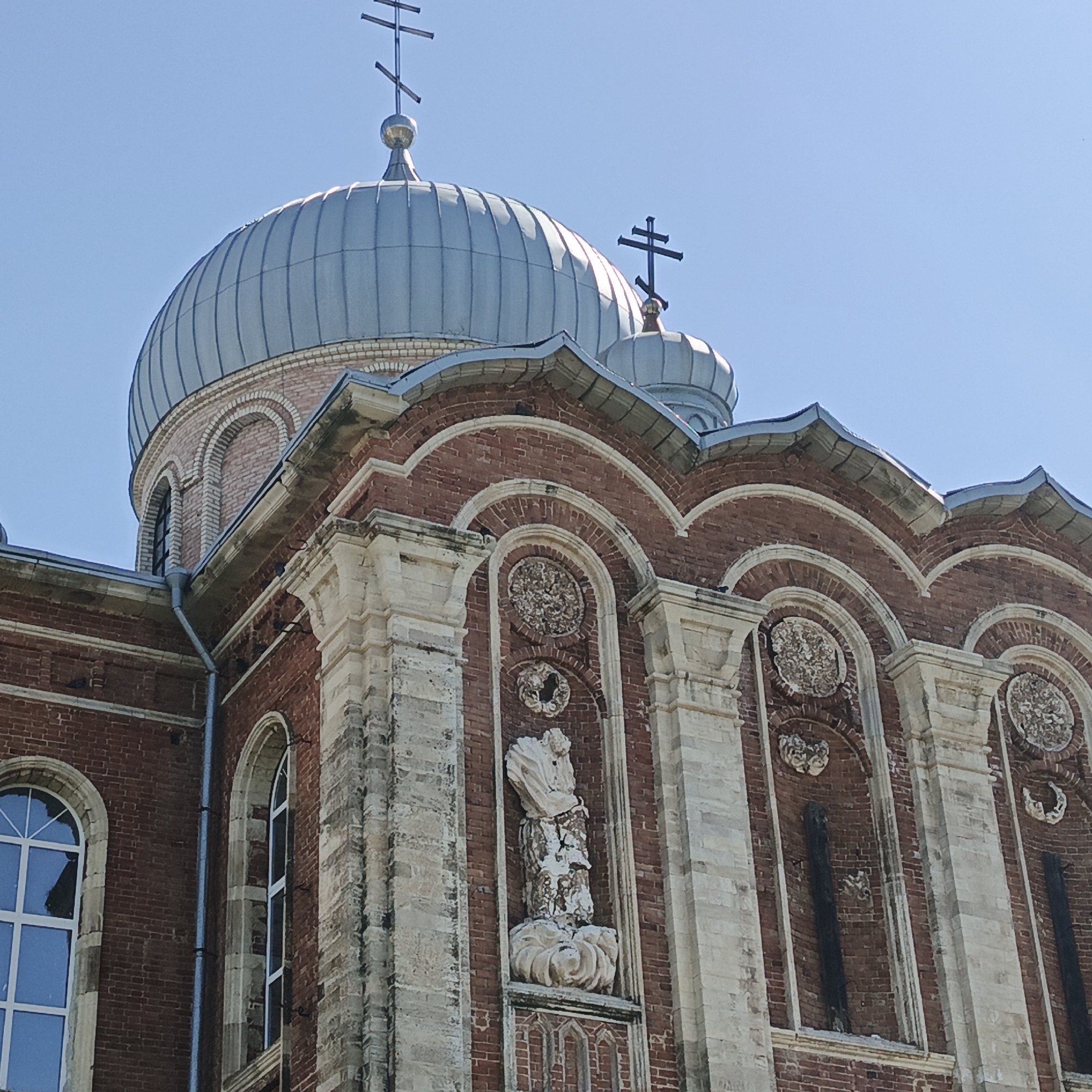 The image shows an exterior view of the Saint Nicholas Church in Tugustemir, Russia. The church features a prominent, ribbed, grey dome topped with a cross. The building itself is constructed from red brick with decorative white stone elements. There are arched windows and niches along the facade. One niche contains a weathered white stone sculpture of a figure. The sky above is clear and blue.